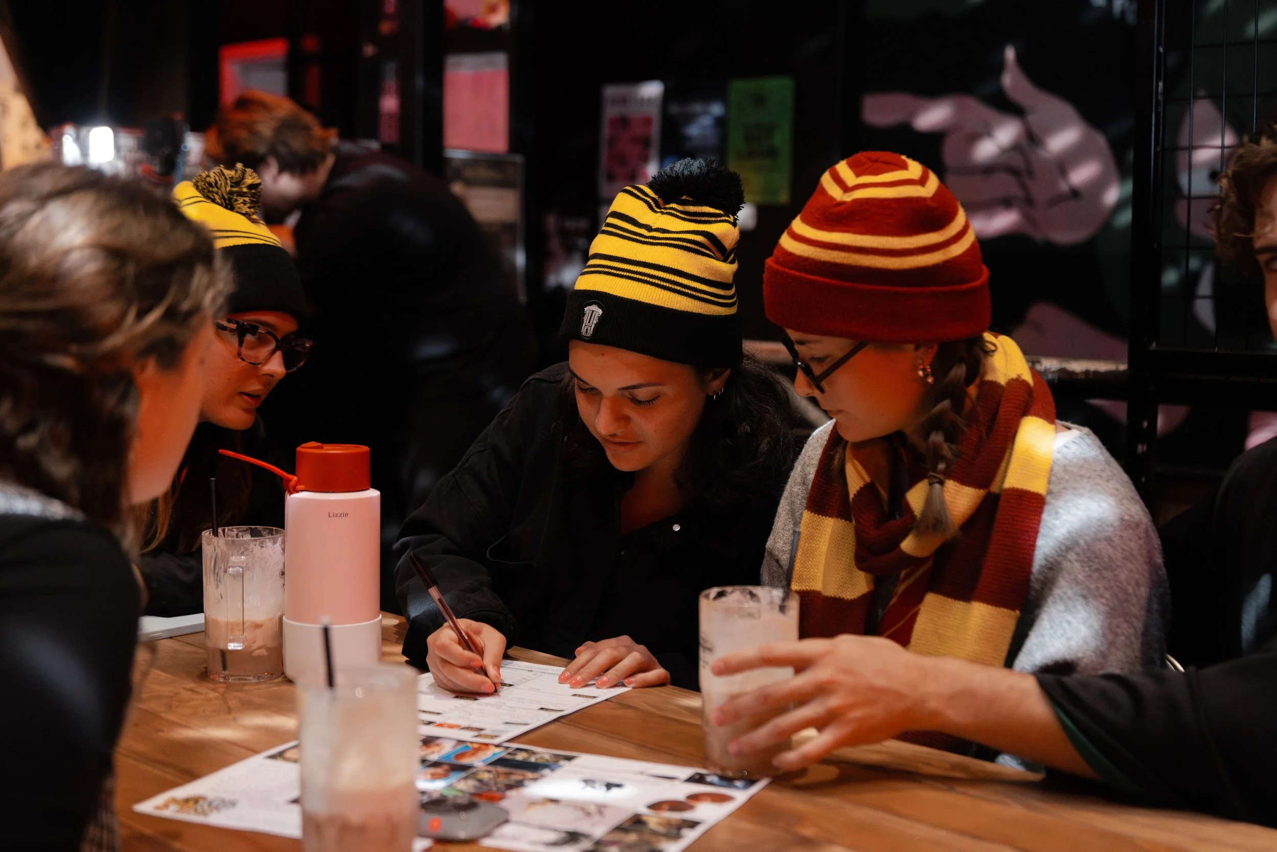 Group of people sitting at a wooden table, wearing winter hats and scarves, looking at papers and writing, with drinks on the table, in a dimly lit indoor setting.