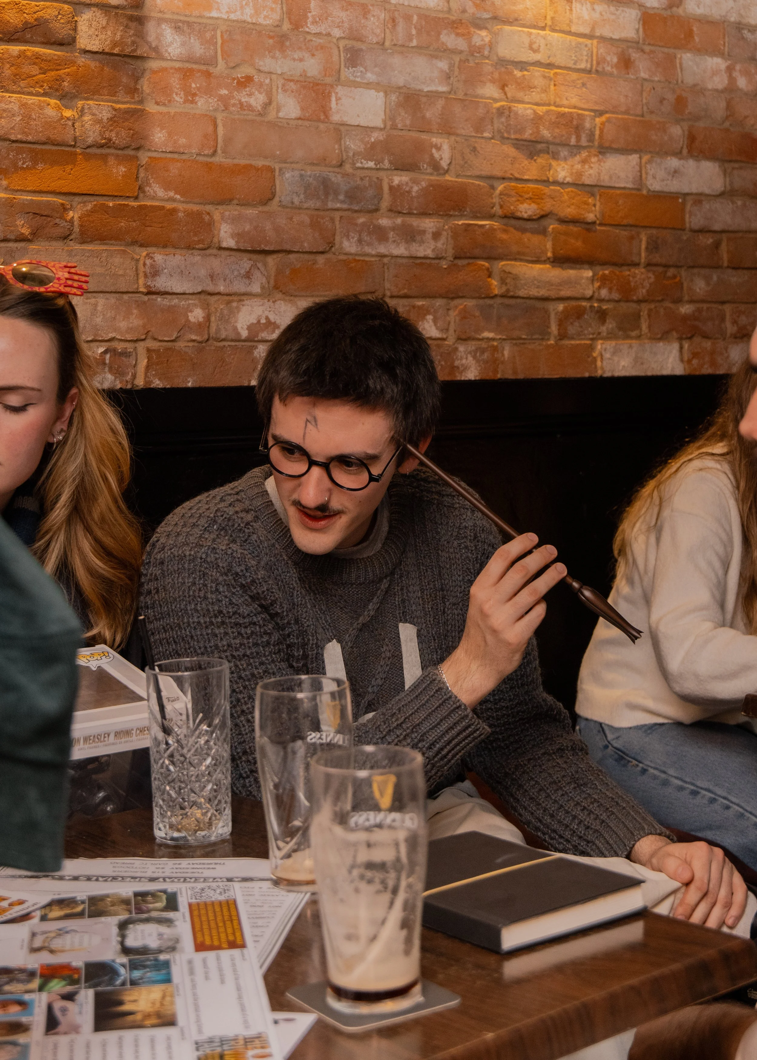 People sitting at a social gathering, with a brick wall in the background. A man with glasses and a painted lightning bolt on his face is holding a cigarette, with drinks and papers on the table in front of him.