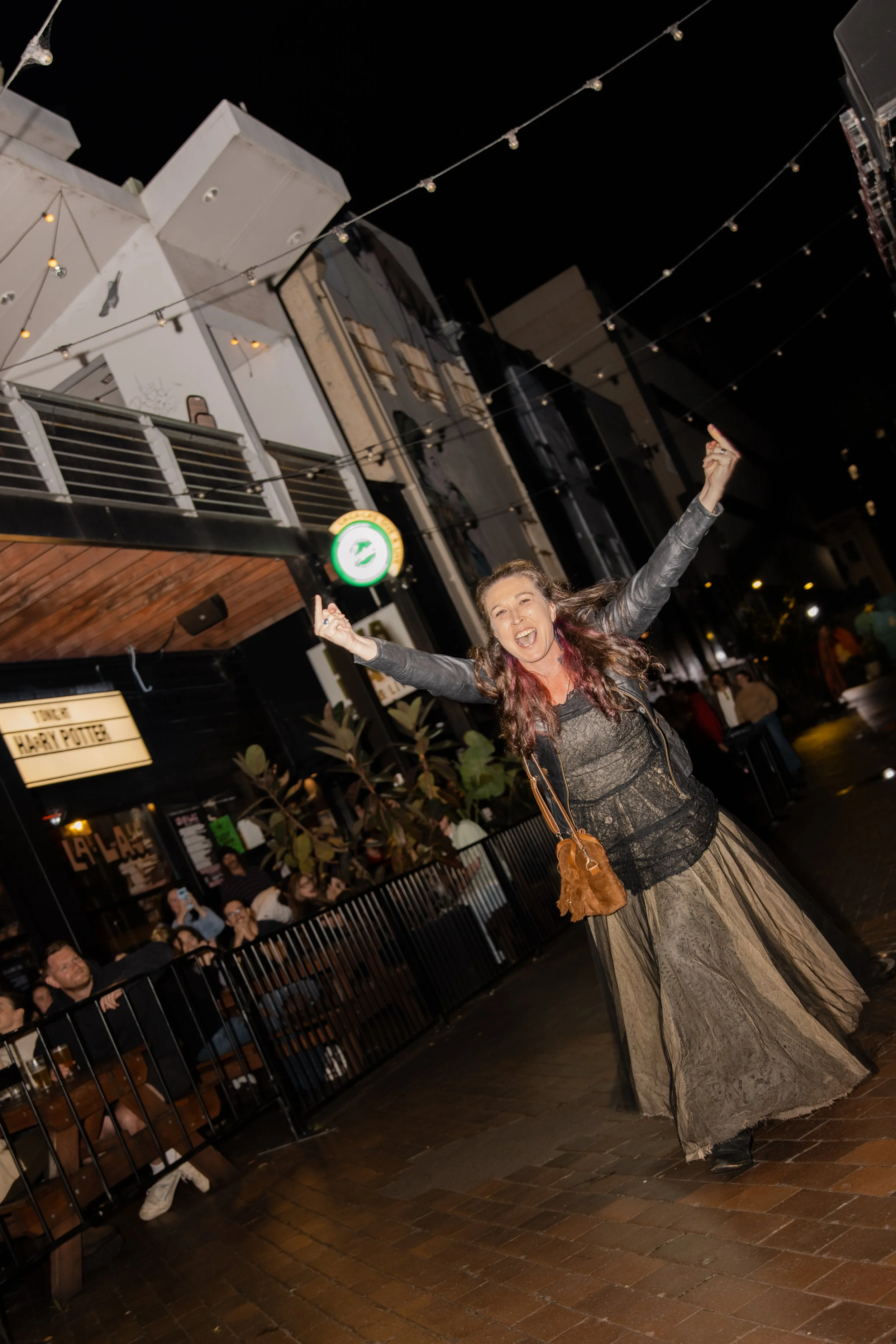 A woman with long hair wearing a gray dress and a black leather jacket is dancing or celebrating outside at night. She has her arms raised and is smiling widely. There are string lights above and a crowd of people watching her near a metal fence, wit