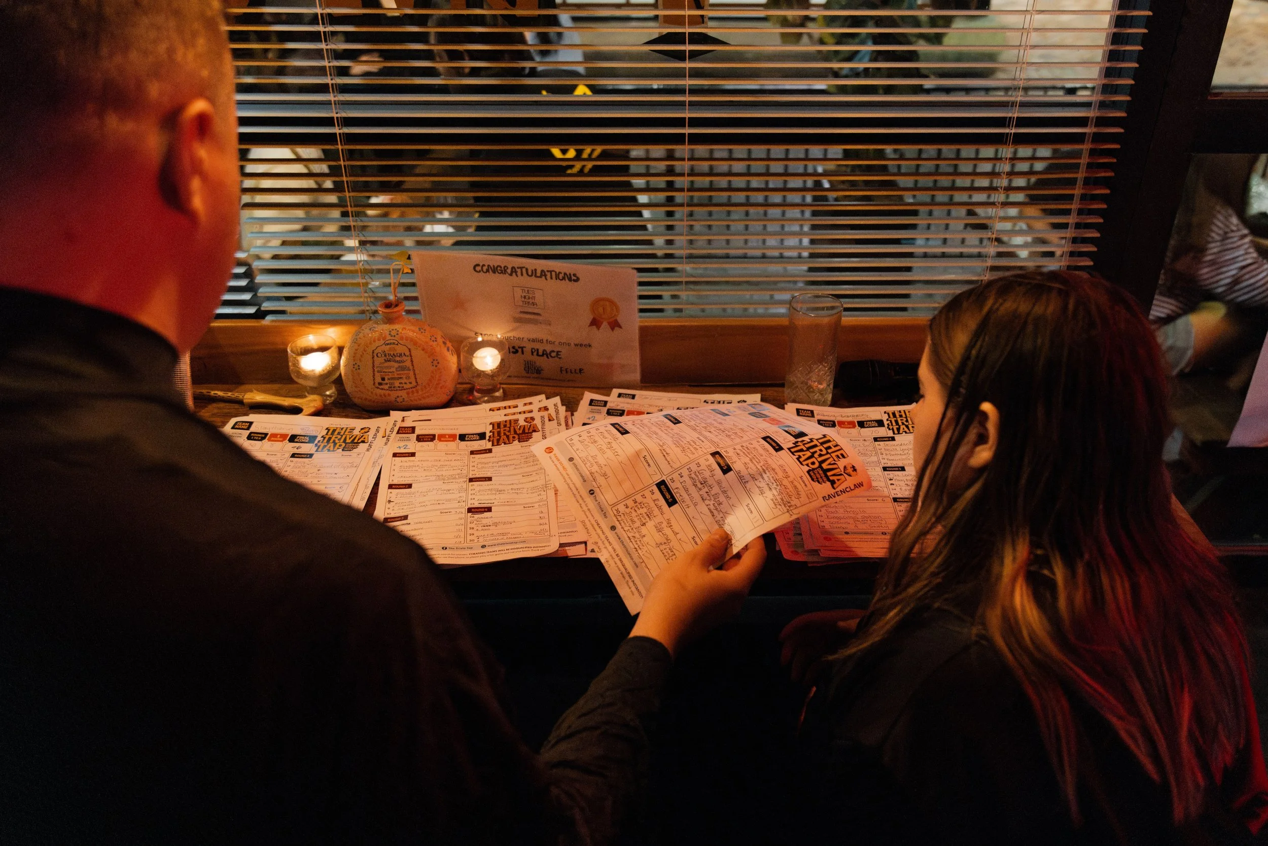People sitting at a table playing trivia, with windows covered by blinds and a sign celebrating a first-place win.