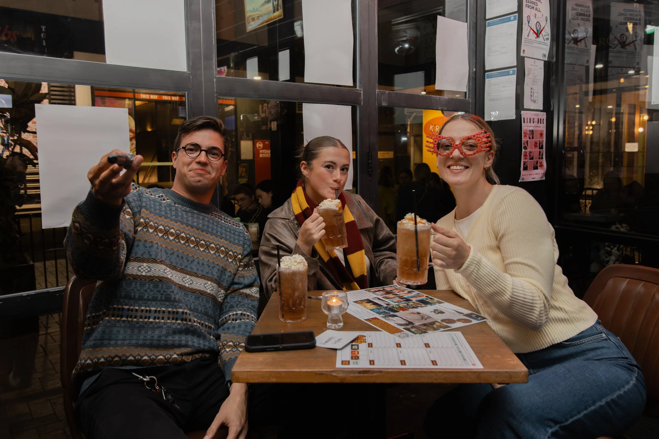 Three friends enjoying drinks at a restaurant table. One person is smiling widely with special glasses, another is sipping a beverage through a straw, and the third is flexing their arm while holding a drink. They are seated indoors with a busy backg