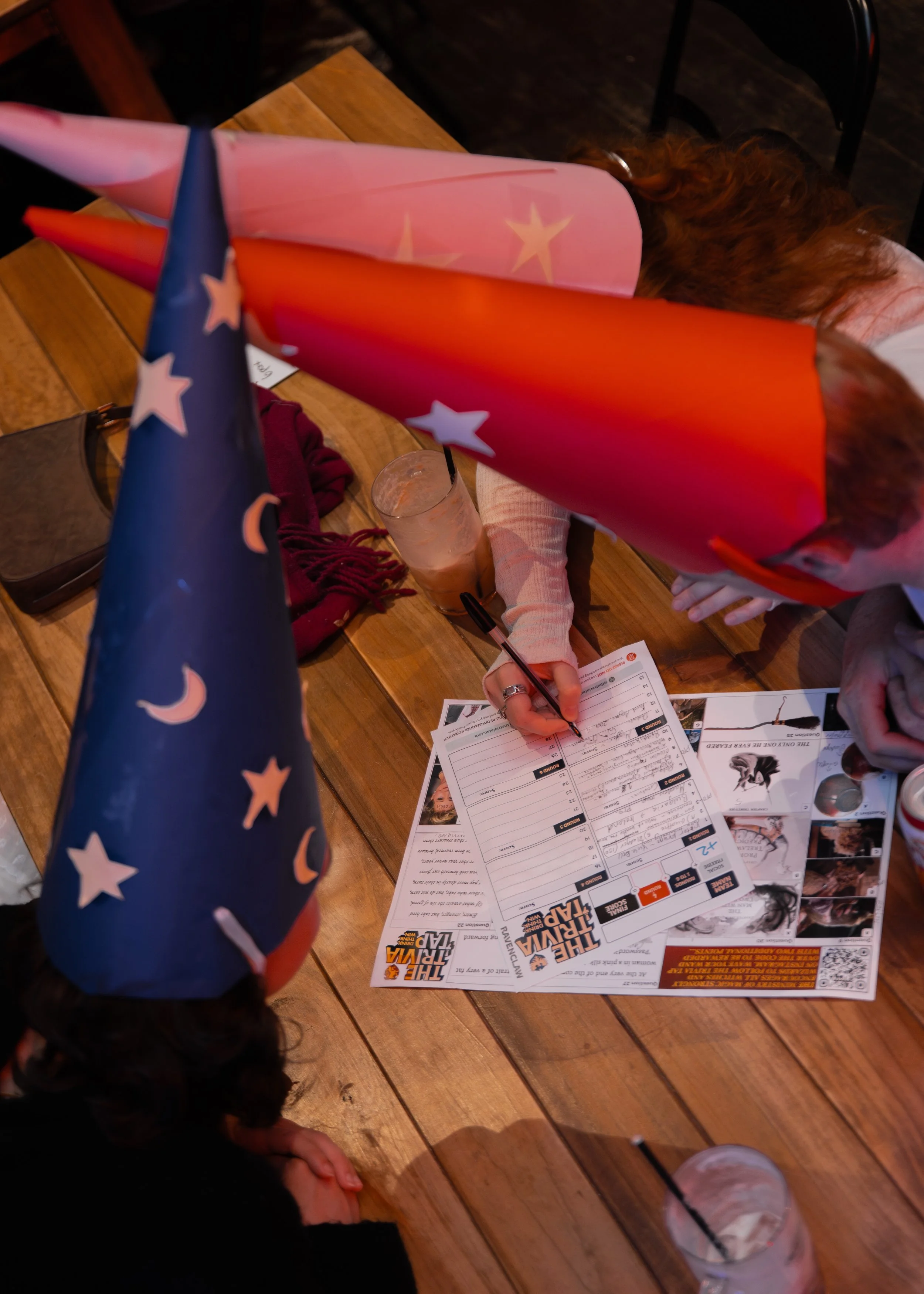 Child wearing a star-spangled party hat, working on a score sheet for a game of The Iron Trap, with beverages and party supplies on a wooden table.