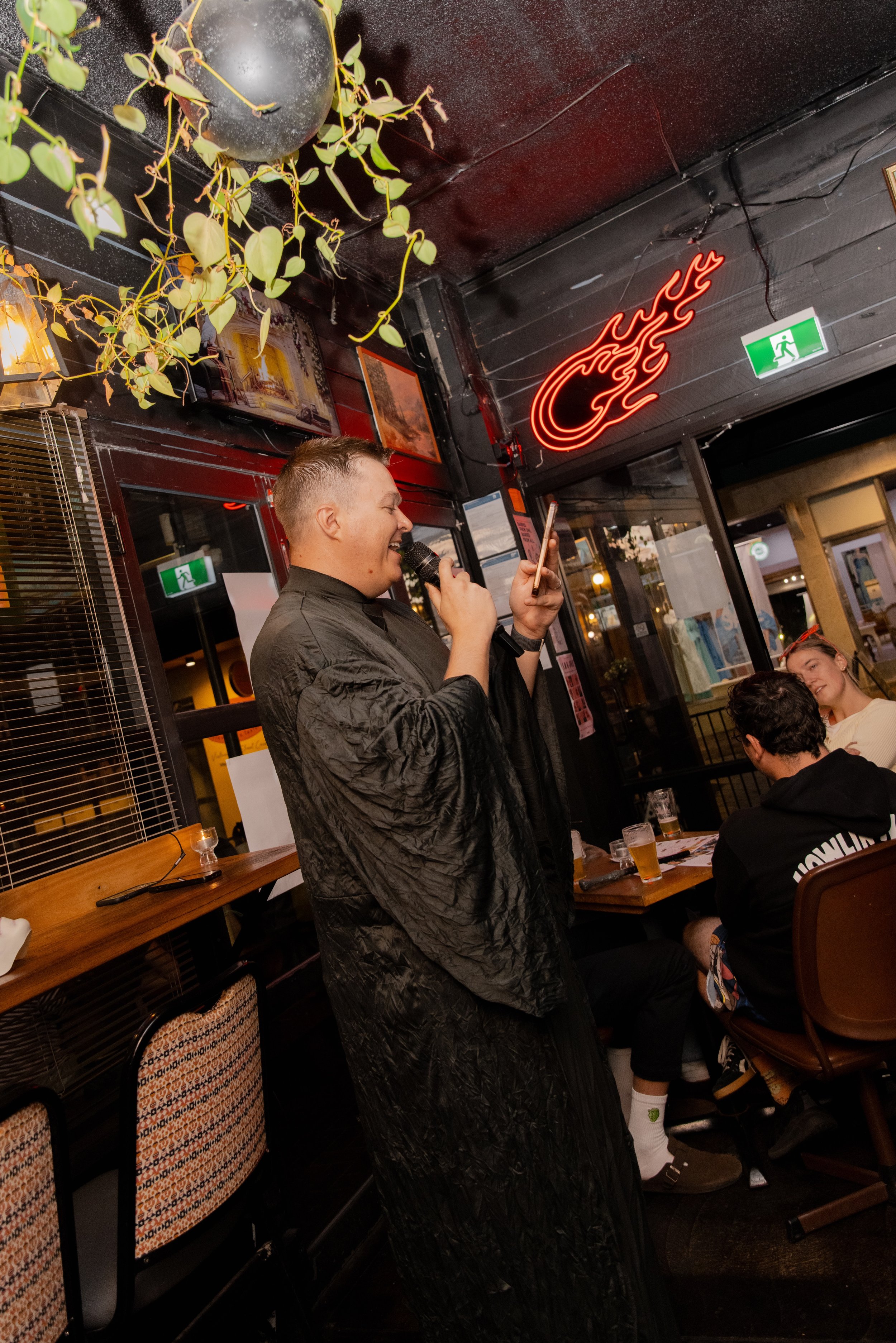 A man with short blond hair wearing a black jacket is speaking or singing into a microphone while looking at his phone in a restaurant or bar. There are other people seated at the table with drinks. The background features a black ceiling, a neon sig