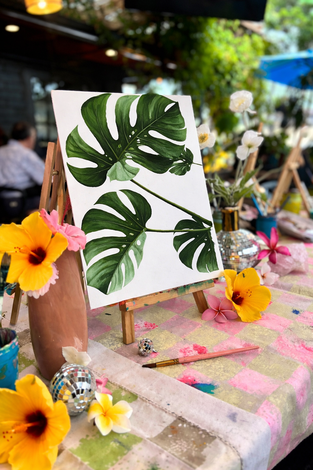 Close-up of a painting of green monstera leaves on a canvas displayed on a small easel, with colorful flowers and art supplies on a checkered table.