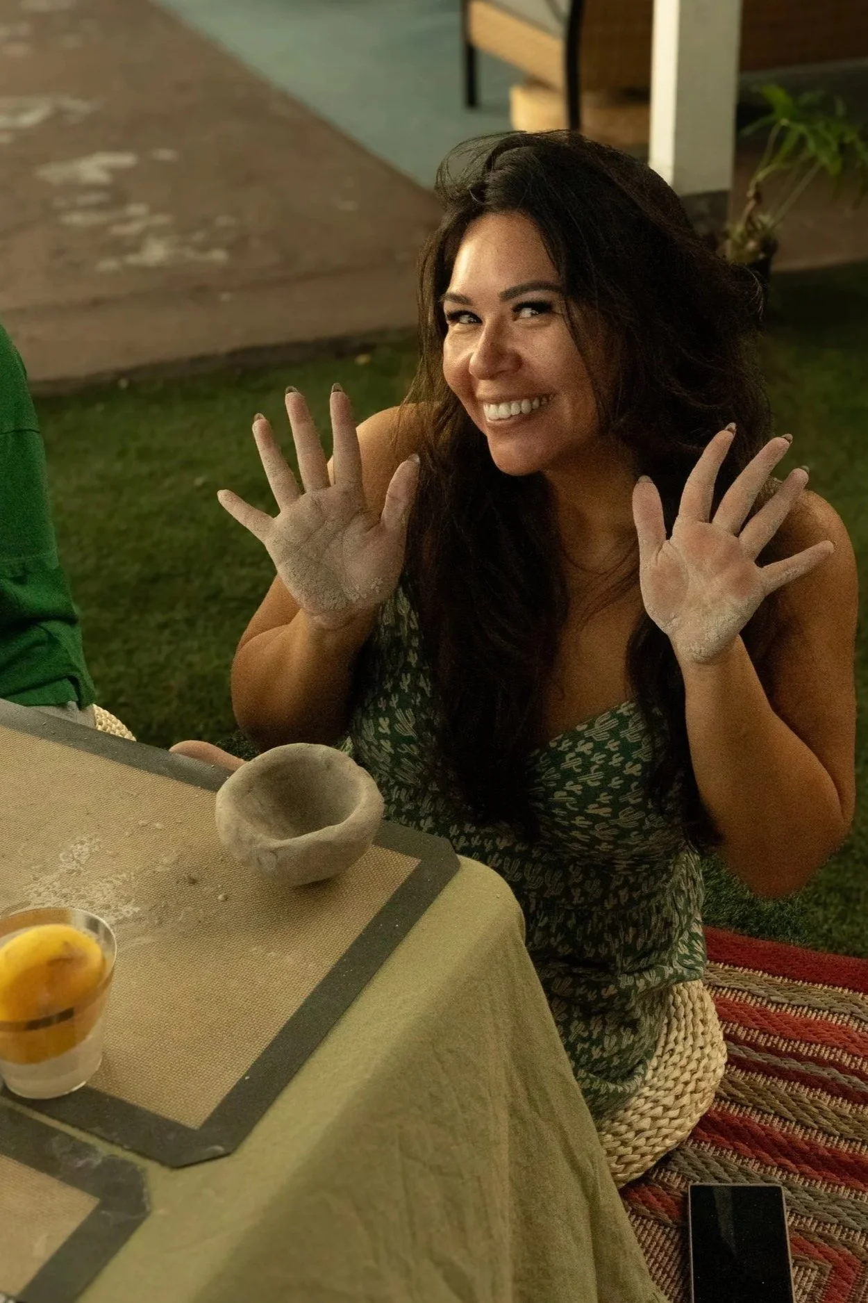 A woman smiling and showing her hands covered in white powder, sitting at a table with a bowl and a glass of yellow liquid, possibly involved in a baking or pottery activity.
