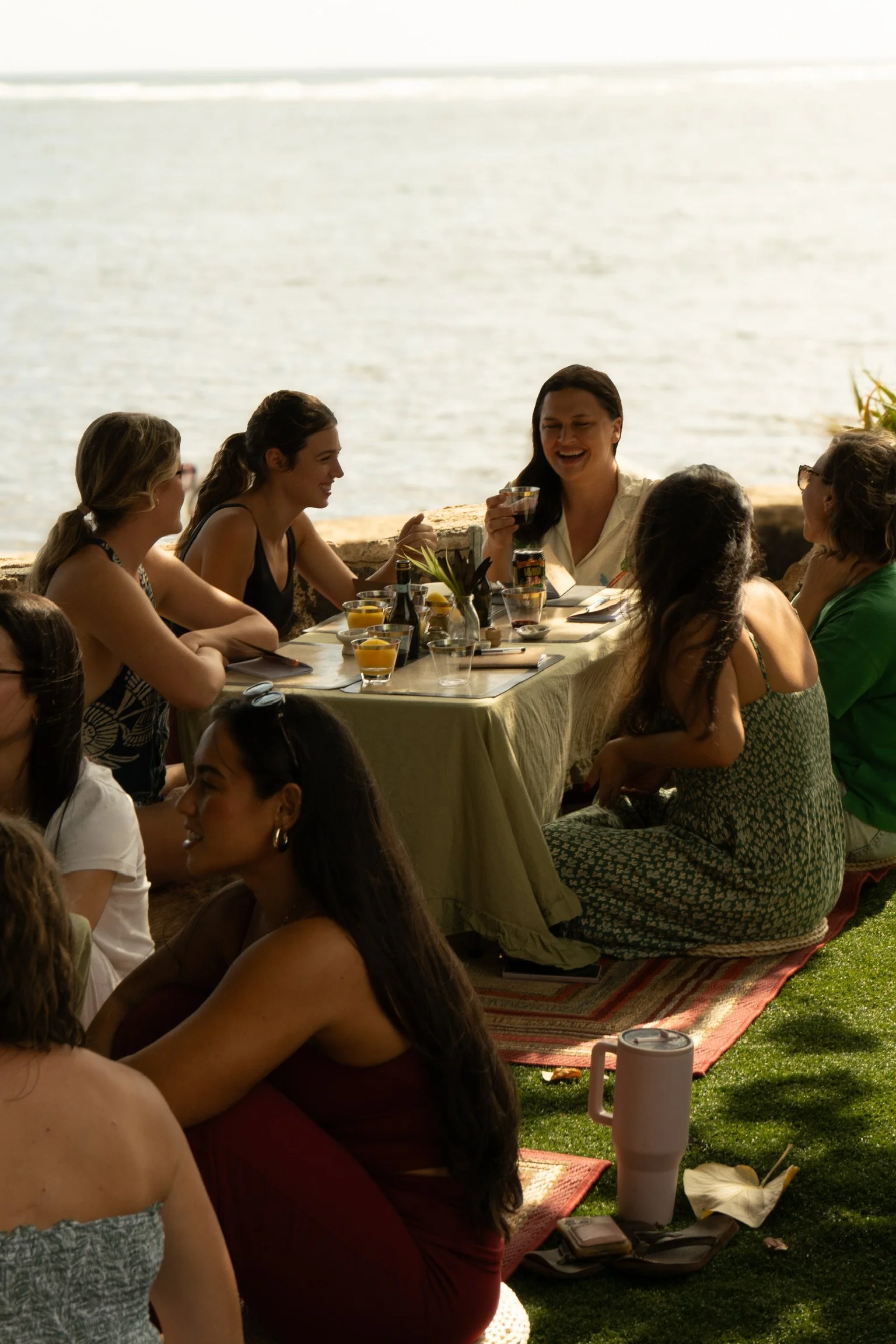 A group of women gathered outdoors by a body of water, enjoying a meal together at a table with drinks and a tablecloth, some sitting on rugs on the grass.