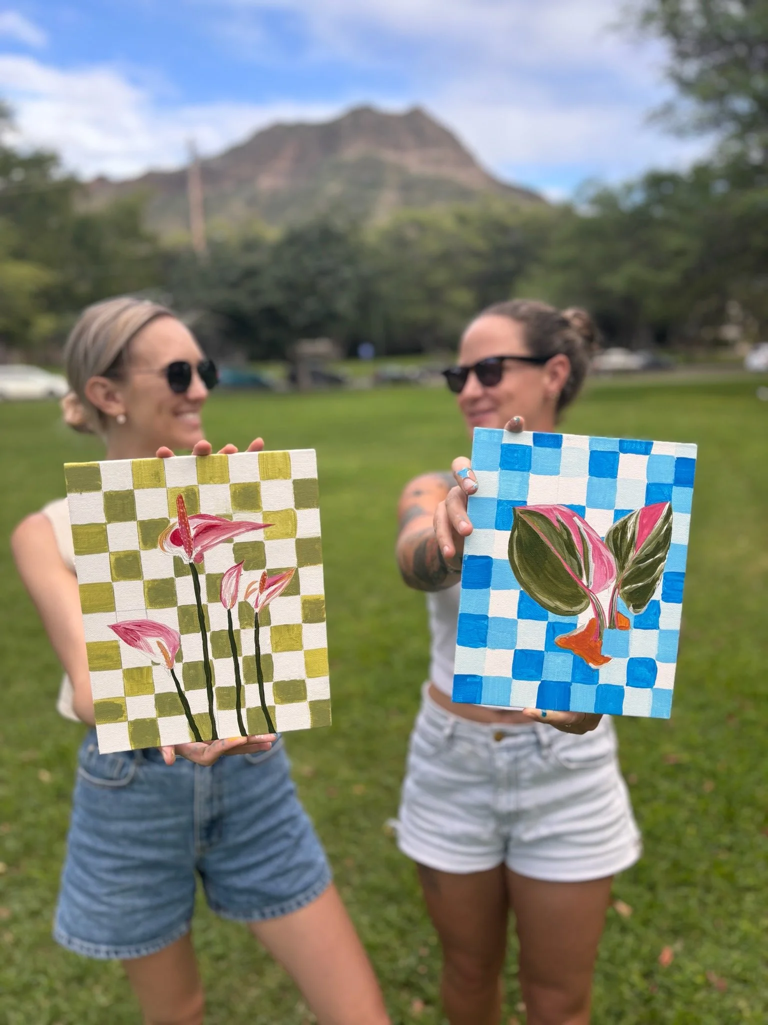 Two students hold up their finished paintings, a pink anthurium on green checks and a pink-leaf plant on blue checks, at an outdoor paint class on Oʻahu.
