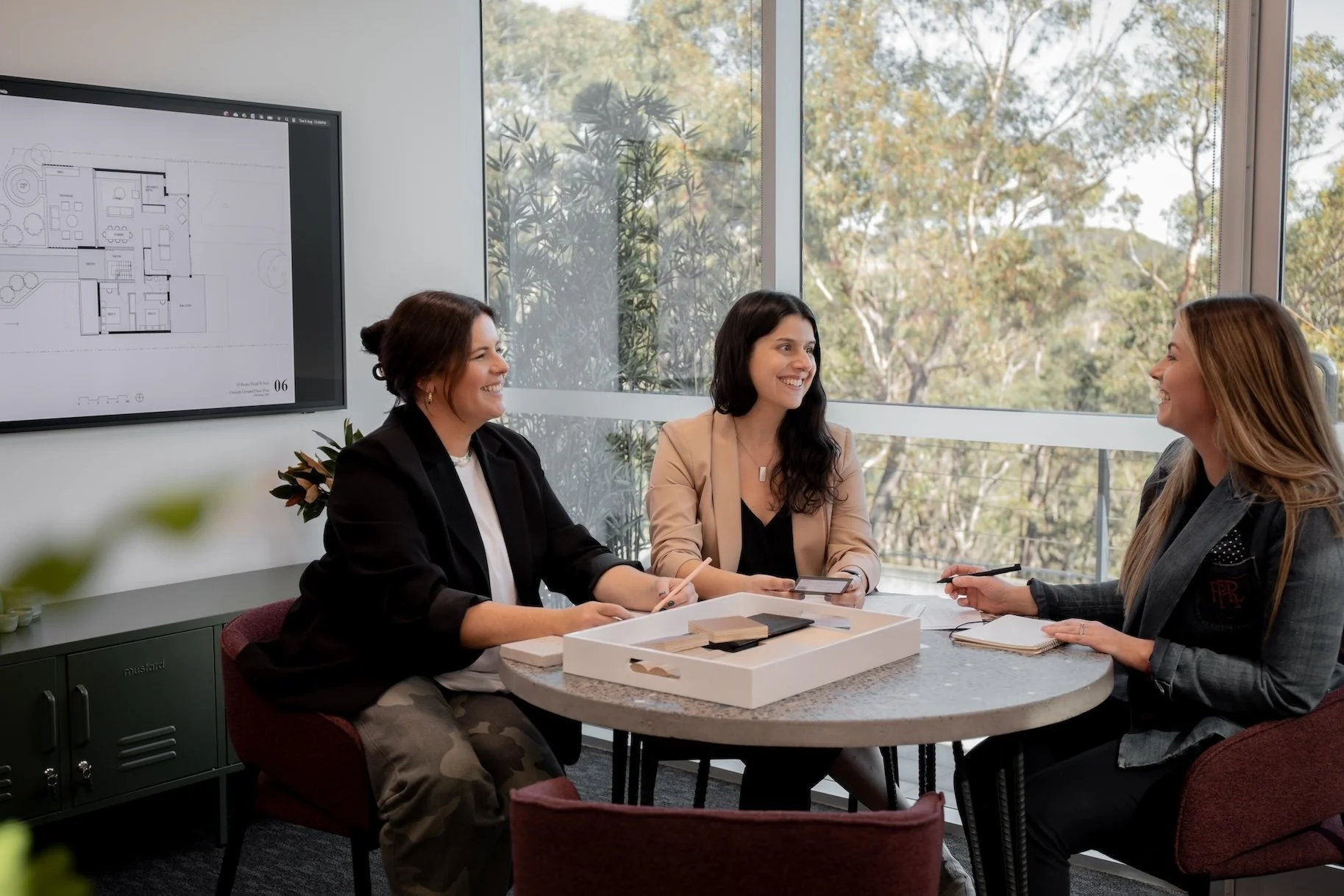 Three women sitting around a round table in a bright office, having a discussion. One woman has a tablet, another has a notepad, and a third has a pen and notebook. A large window shows trees outside, and a monitor with a floor plan is on the wall.