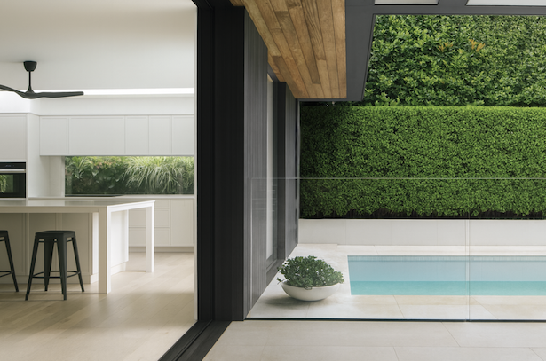 Modern kitchen with white cabinets, a kitchen island, black bar stools, and a ceiling fan, adjacent to a backyard with a swimming pool and a green hedge.