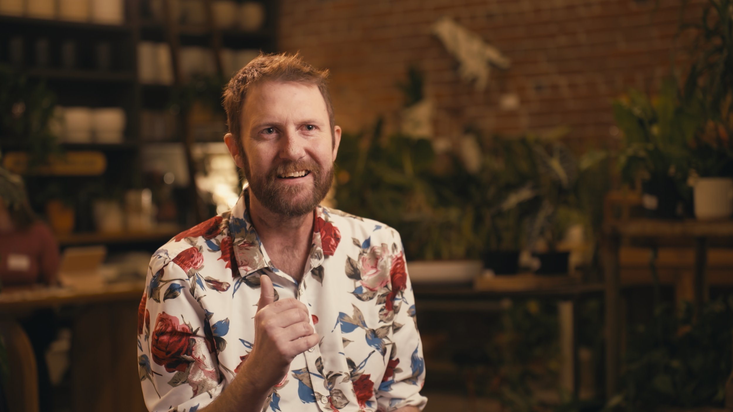 Man wearing a floral shirt, sitting indoors with plants and shelves in the background — captured in a high-quality Sacramento video.