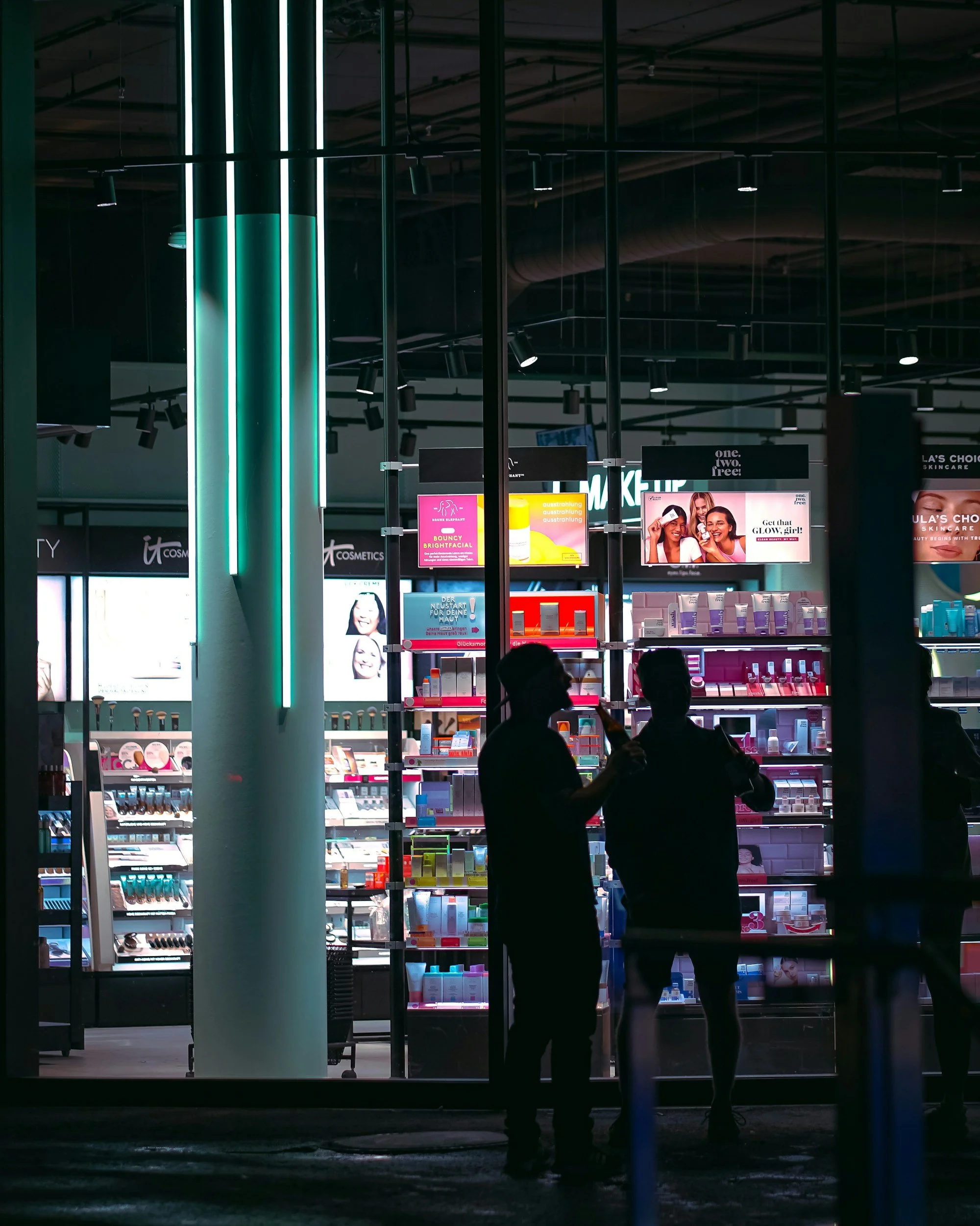 A nighttime view of a store with illuminated displays and advertisements visible inside through large glass windows. Two silhouetted people are standing outside, facing the store, with one person appearing to speak or gesturing.