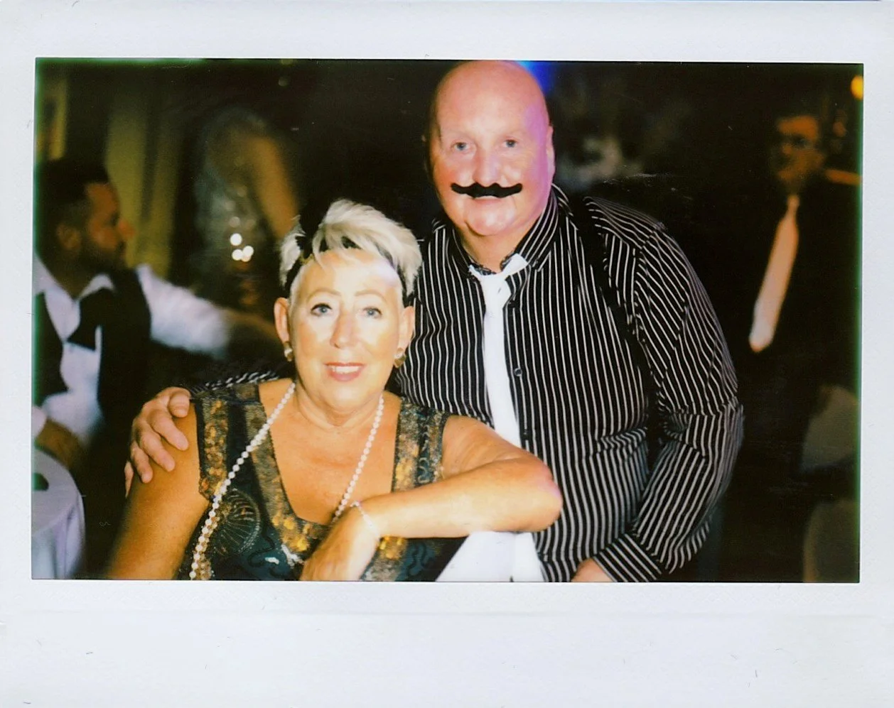 Photobooth Hire Scotland – A woman and a man at a social event, with the man wearing a striped shirt and a fake moustache, and the woman wearing a fancy dress and pearl necklace. The man has his arm around the woman and they are smiling.