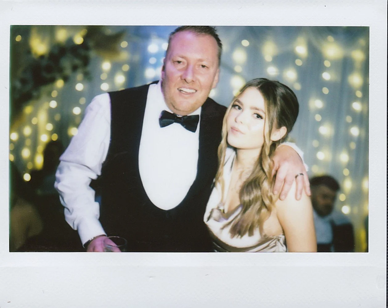 Photobooth Hire Scotland – Luxury Vintage Polaroid Experience | Vintage Instant Photographer A man and a woman dressed in formal attire pose together at an event with decorative lights in the background.