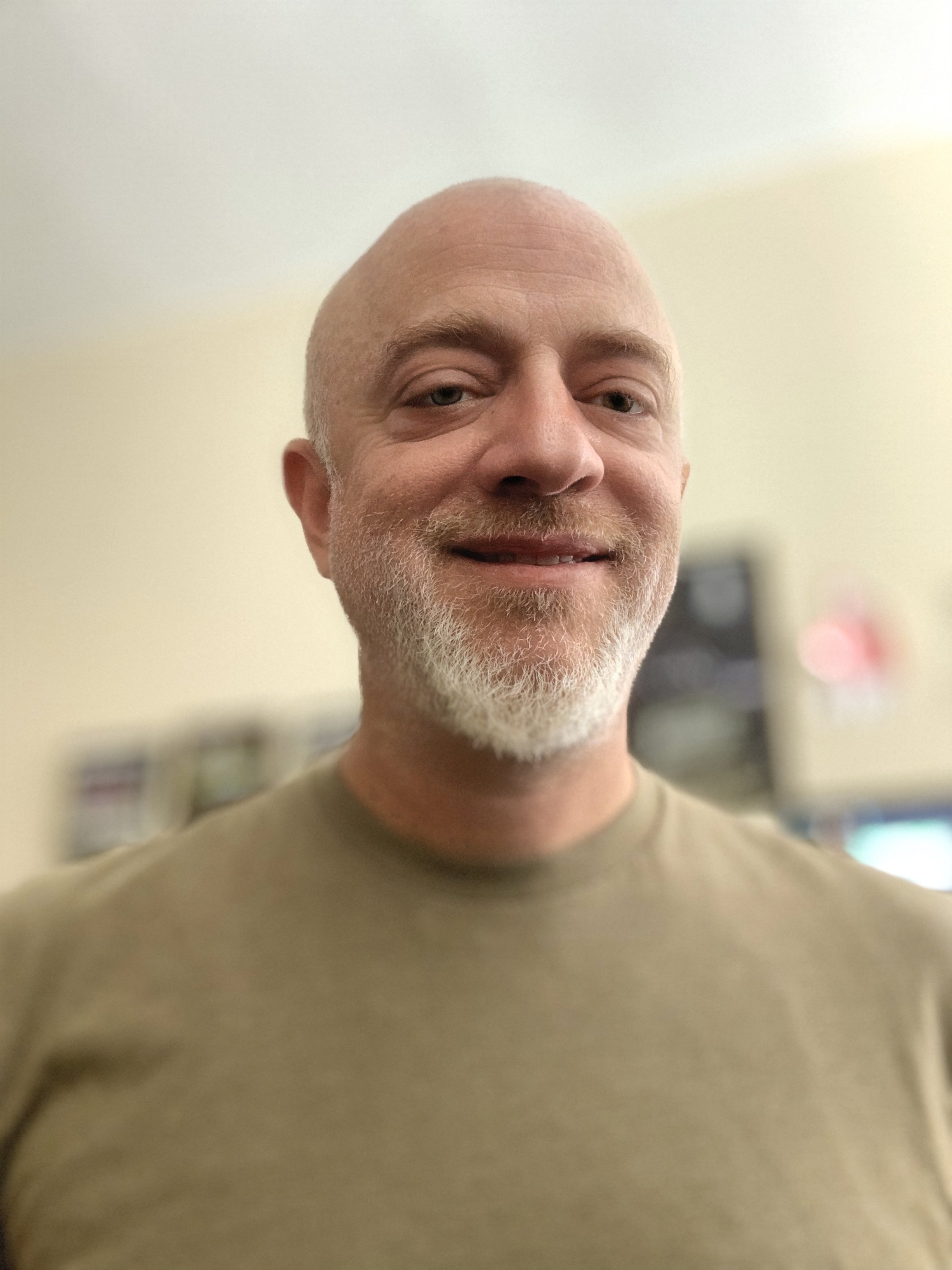 A smiling man with a bald head and white beard, wearing a beige shirt, standing indoors with a blurred background.