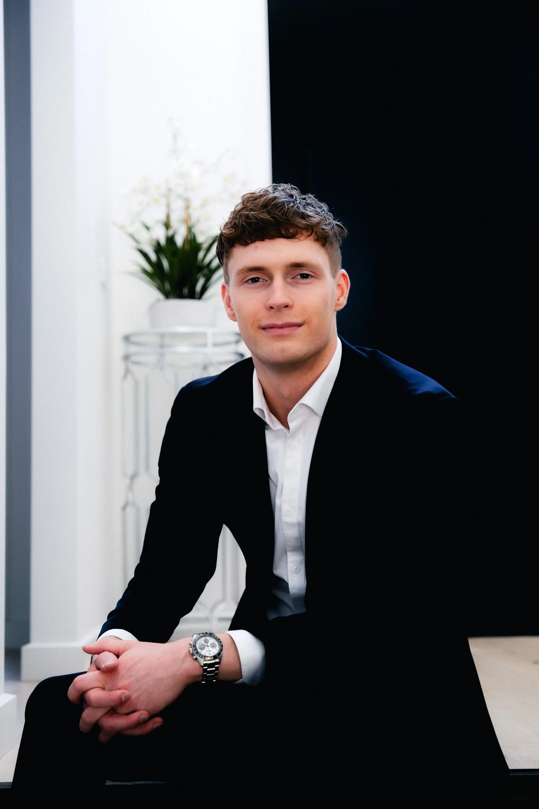 A young man with light skin, short curly hair, wearing a black suit and white shirt, sitting with his hands clasped, in a modern indoor setting with a white wall, a potted plant, and a black partition.