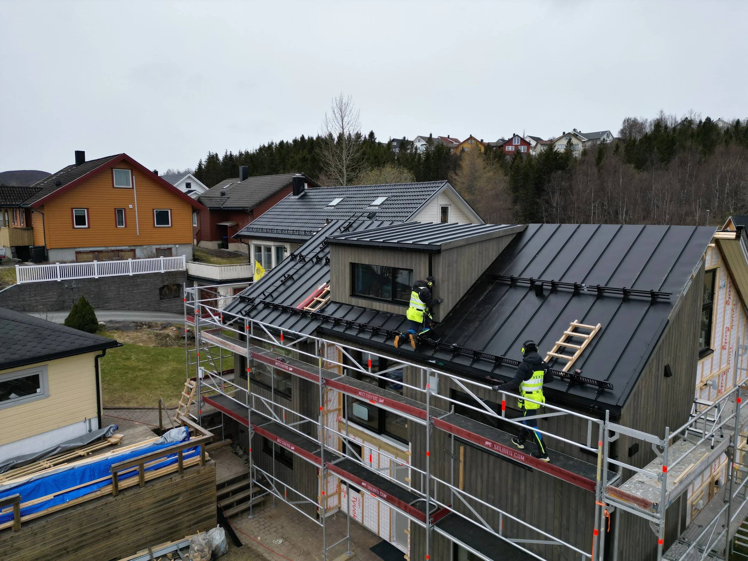To personer i refleksvest arbeider på taket på enebolig. Stillas er satt opp rundt hele huset.