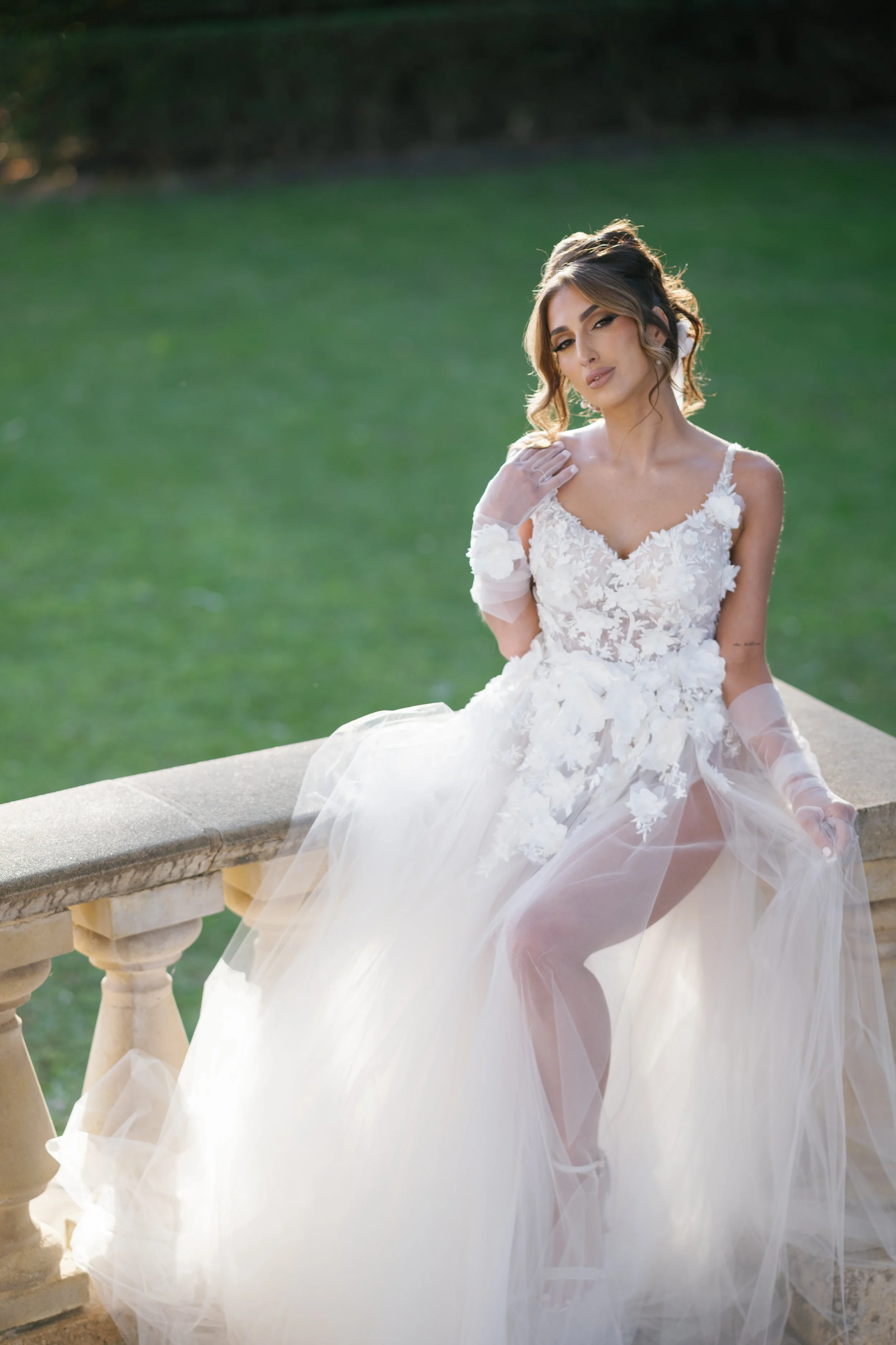 A woman in a wedding dress sitting on a stone railing outdoors with a grassy background.