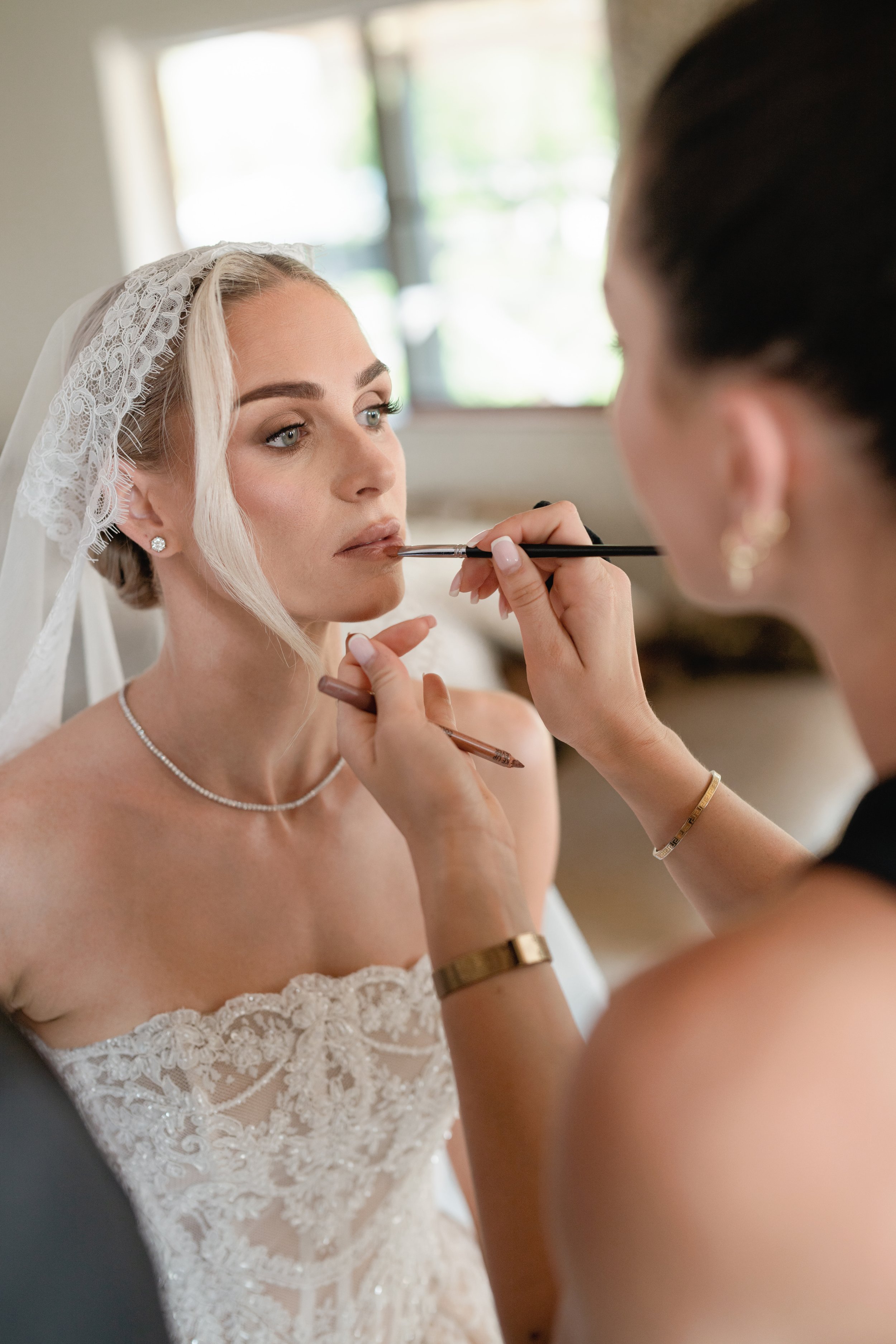 Makeup artist applying lipstick to a bride in a white wedding dress with lace detailing, wearing a lace veil, in a well-lit room.