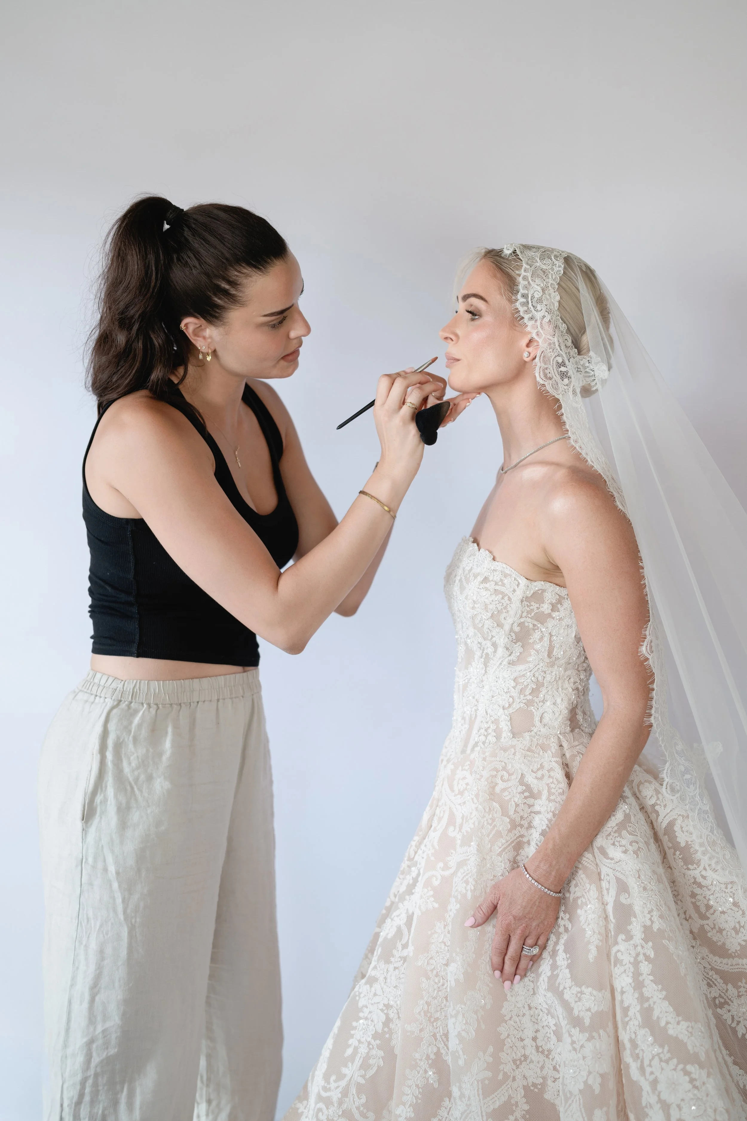 A makeup artist applies makeup to a bride's face, who is wearing a lace wedding gown and veil.