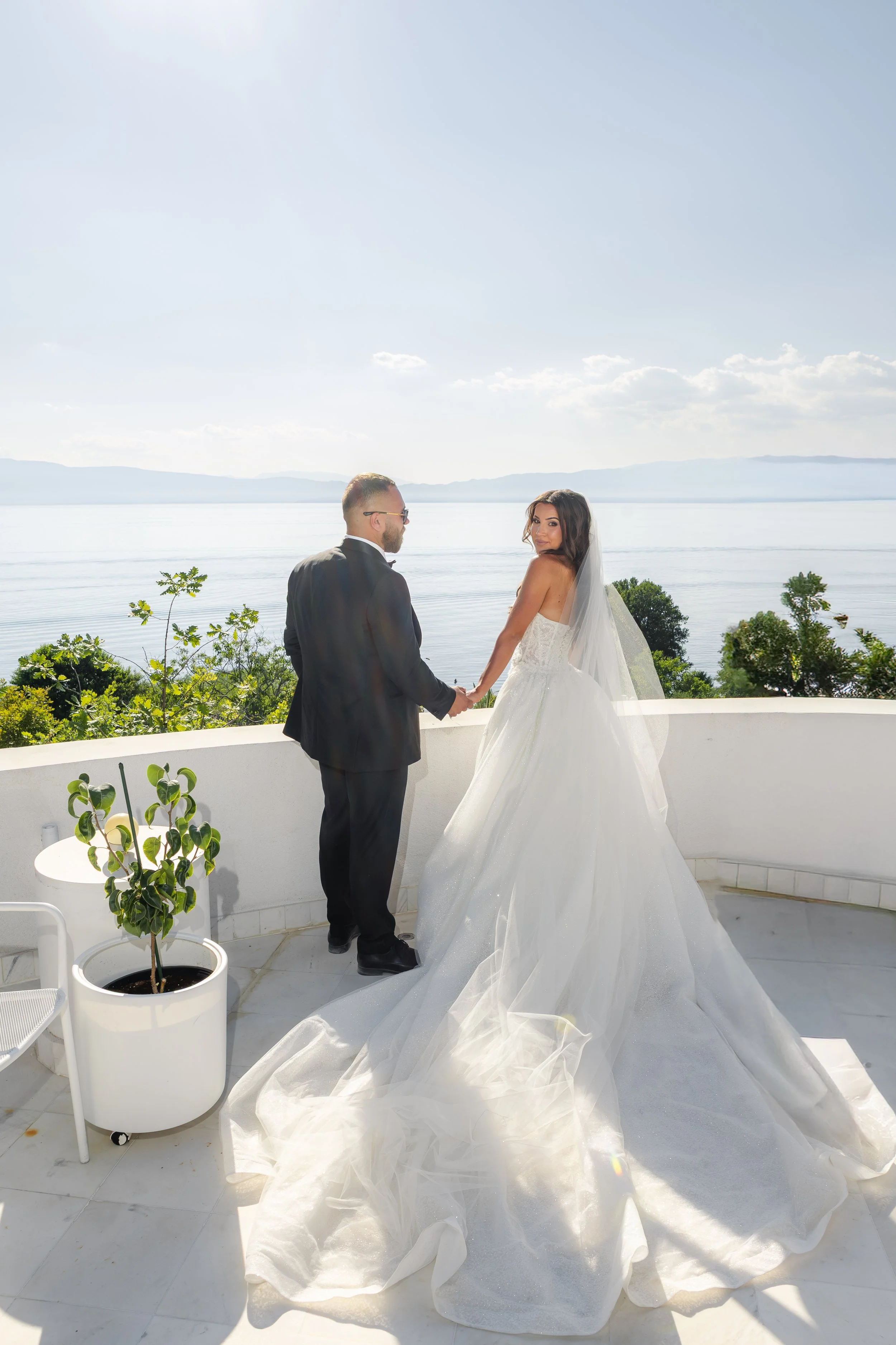 Bride and groom holding hands on a balcony overlooking a lake, with trees and mountains in the background, on a sunny day.