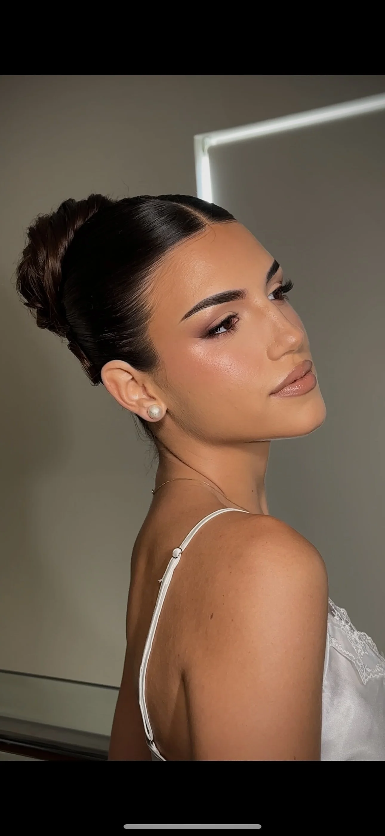A woman with dark hair in a bun, wearing pearl earrings and a white spaghetti strap top, posed against a plain background.