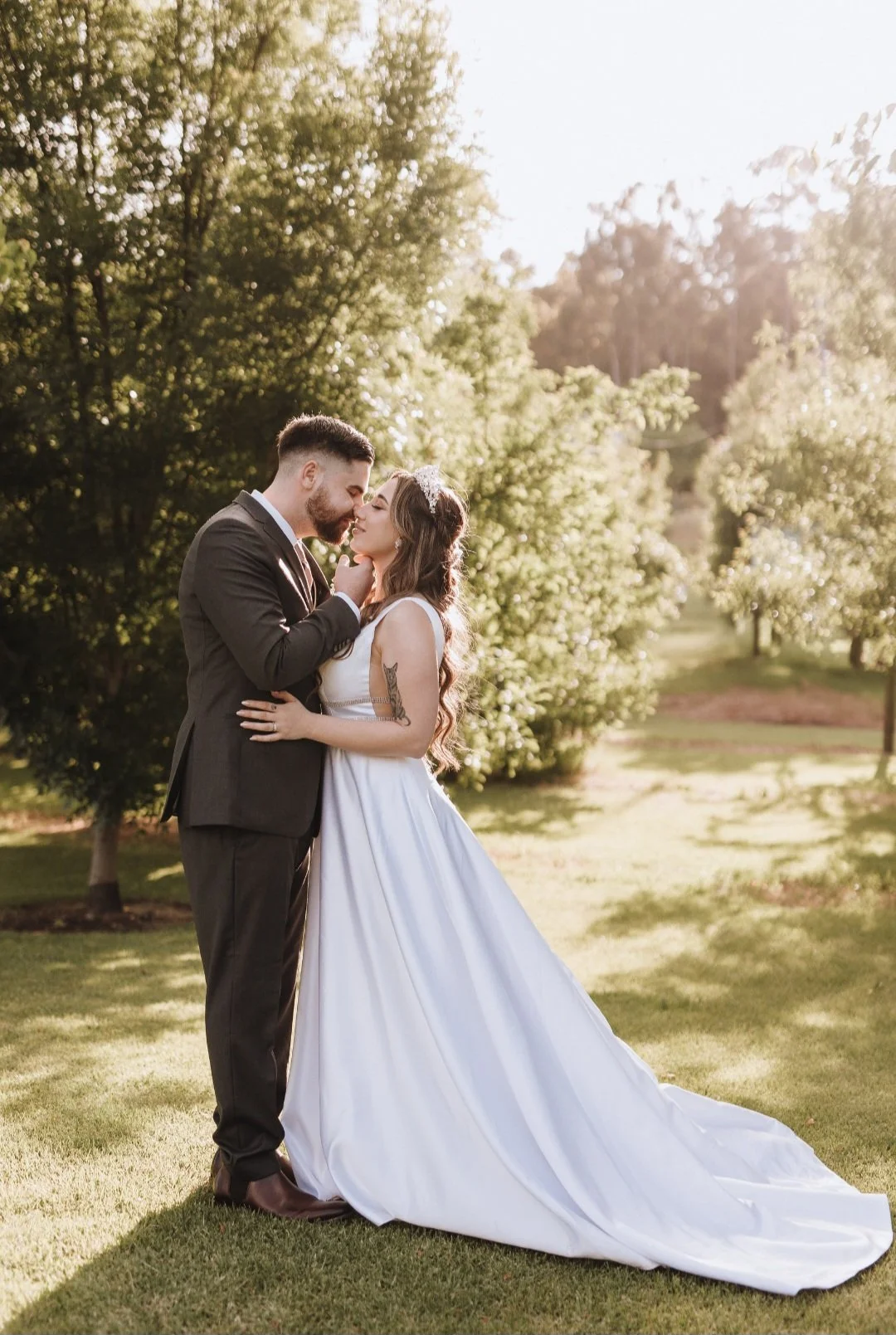 A couple in wedding attire standing close together on a grassy area in a park, with trees and sunlight in the background. The groom is touching the bride's face while they look at each other.