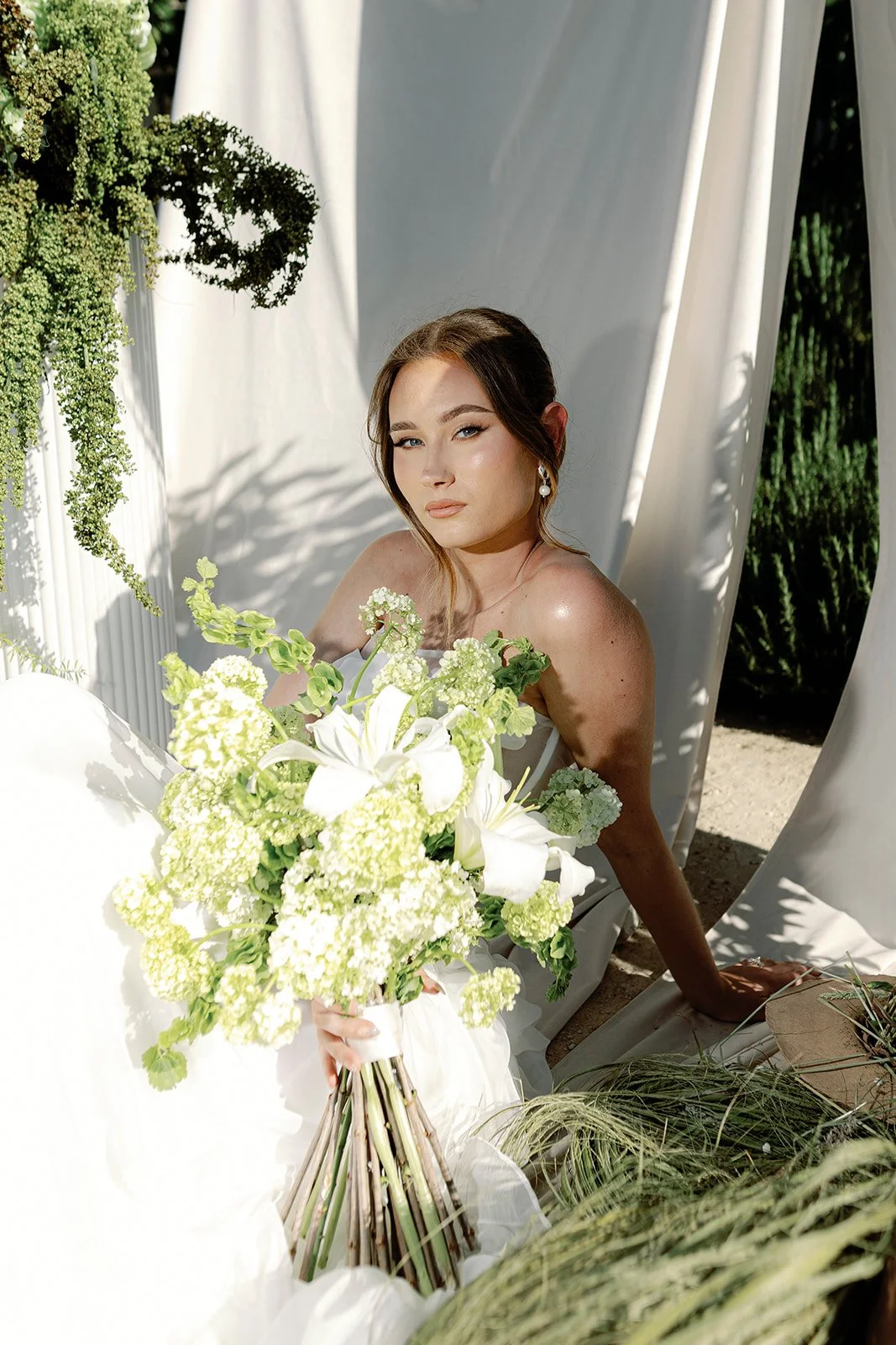 A woman in a white dress holding a large white and green floral bouquet, sitting outdoors with white curtains and greenery in the background.