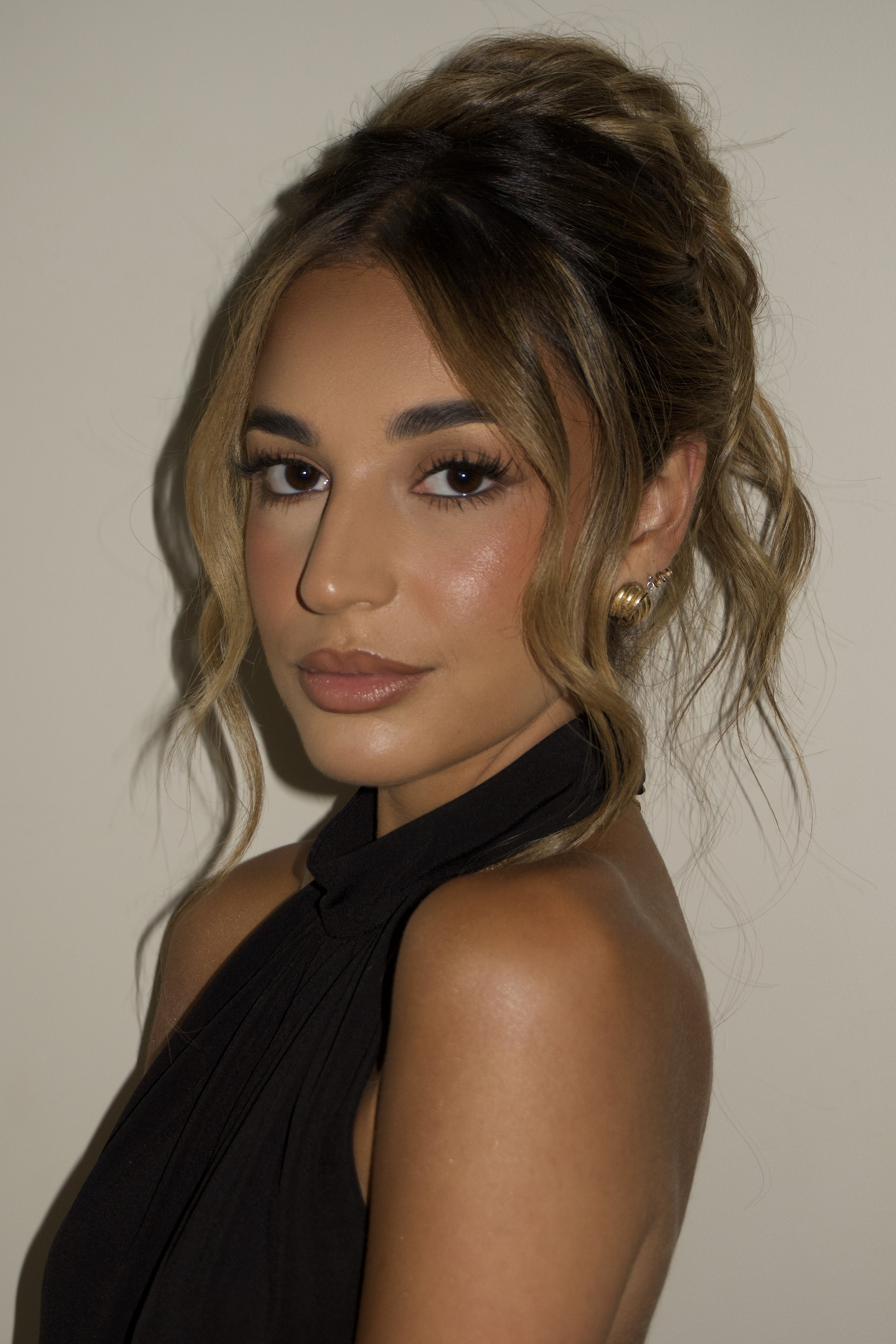 A young woman with wavy blonde and brown hair, wearing a black sleeveless top, has makeup with intense eye makeup and natural lips, and is wearing gold earrings, standing against a plain wall.