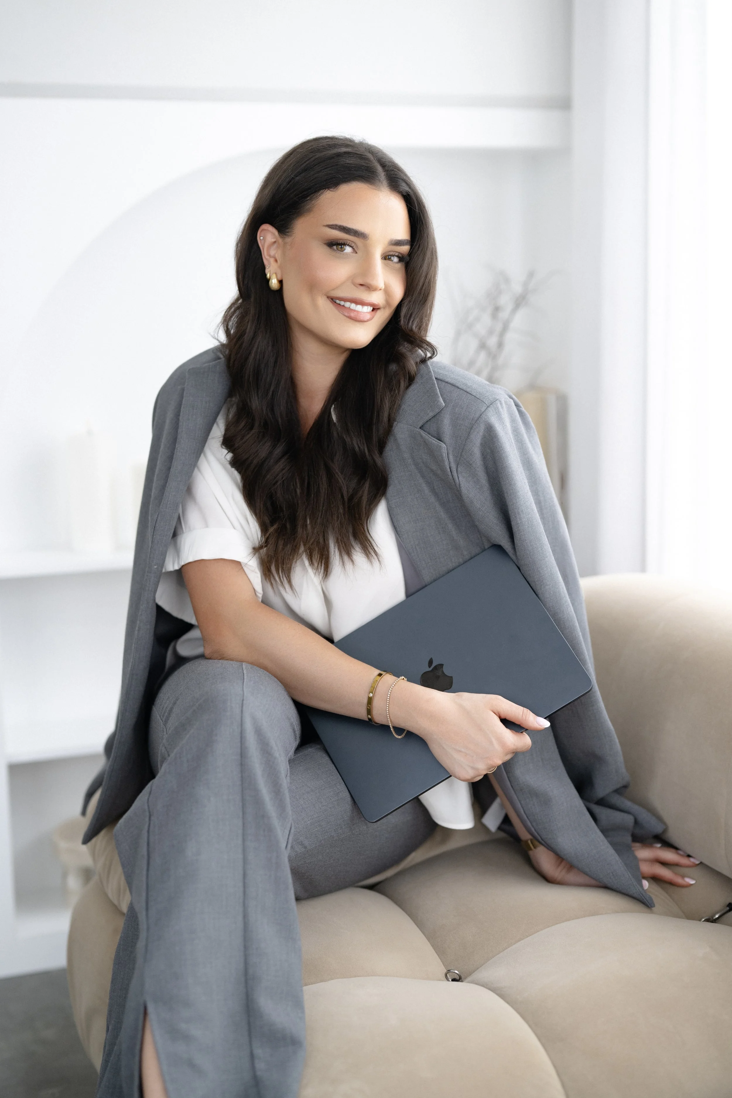 A young woman with long dark hair, wearing a gray blazer and pants, holding a closed MacBook, sitting on a beige couch in a bright, modern room, smiling at the camera.