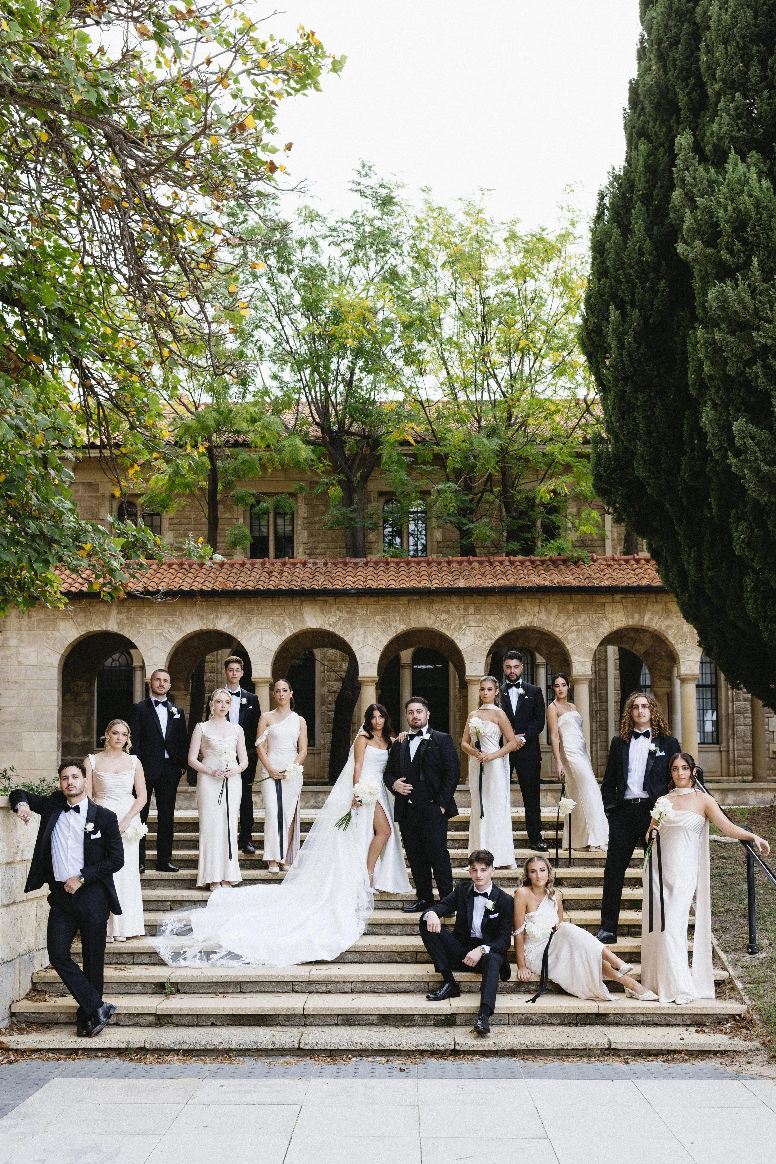 A group of young men and women in formal wedding attire posed on outdoor stone steps in front of an old stone building with arches, trees, and a red-tiled roof.