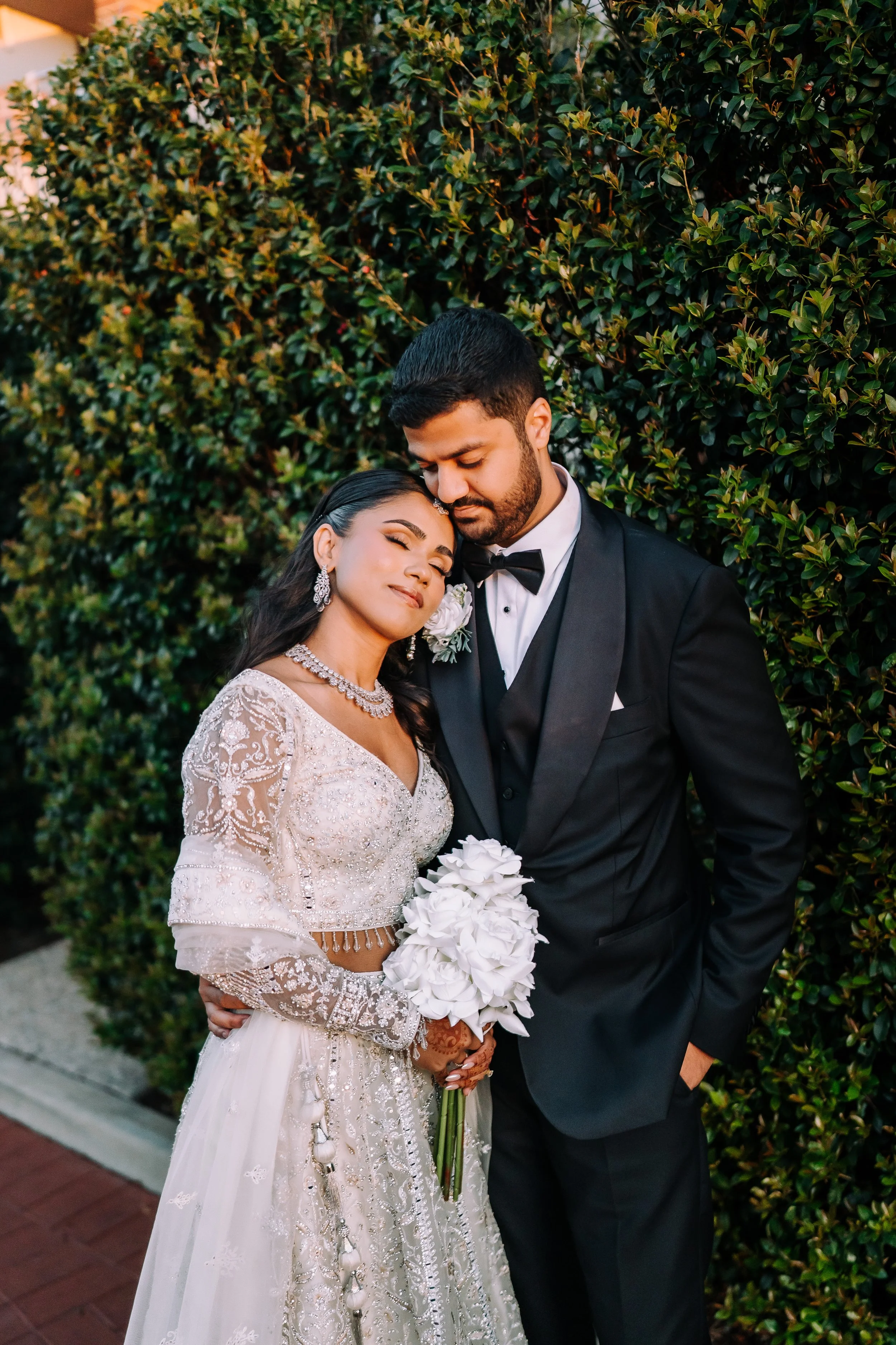 A bride and groom embrace outdoors, the bride holding a bouquet of white flowers and the groom with his hand in his pocket, both dressed in wedding attire against a background of lush greenery.