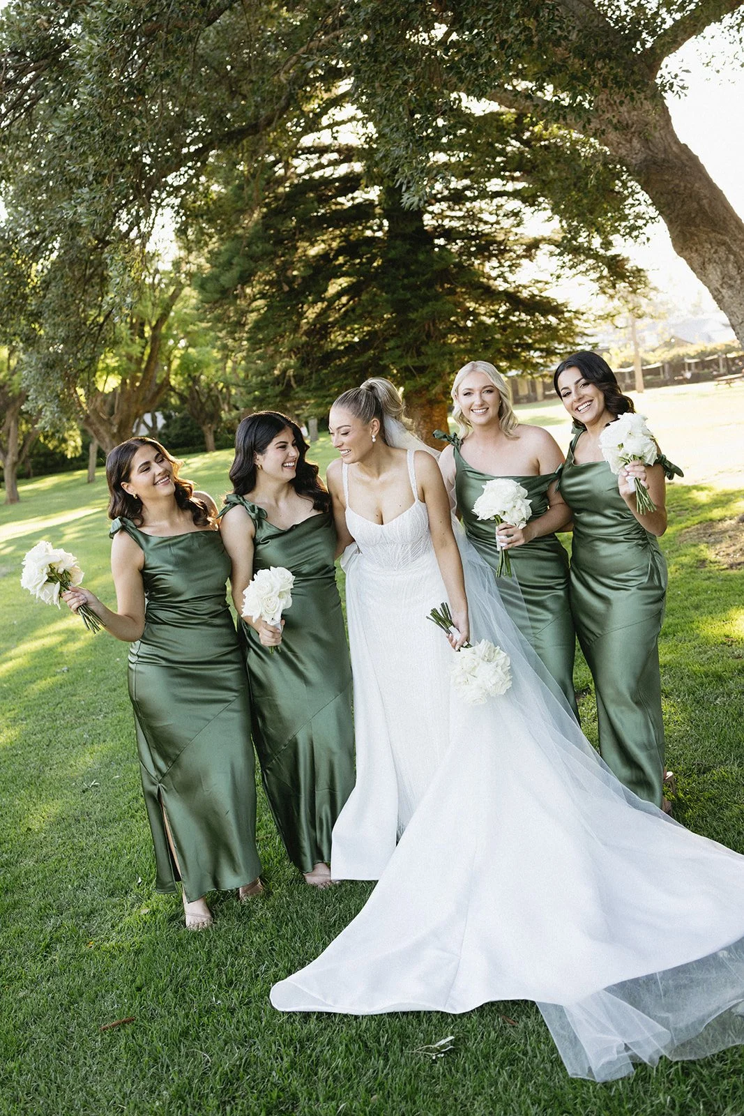 A bride in a white wedding gown surrounded by four bridesmaids in green satin dresses, holding white bouquets, under a large tree in a park during daytime.