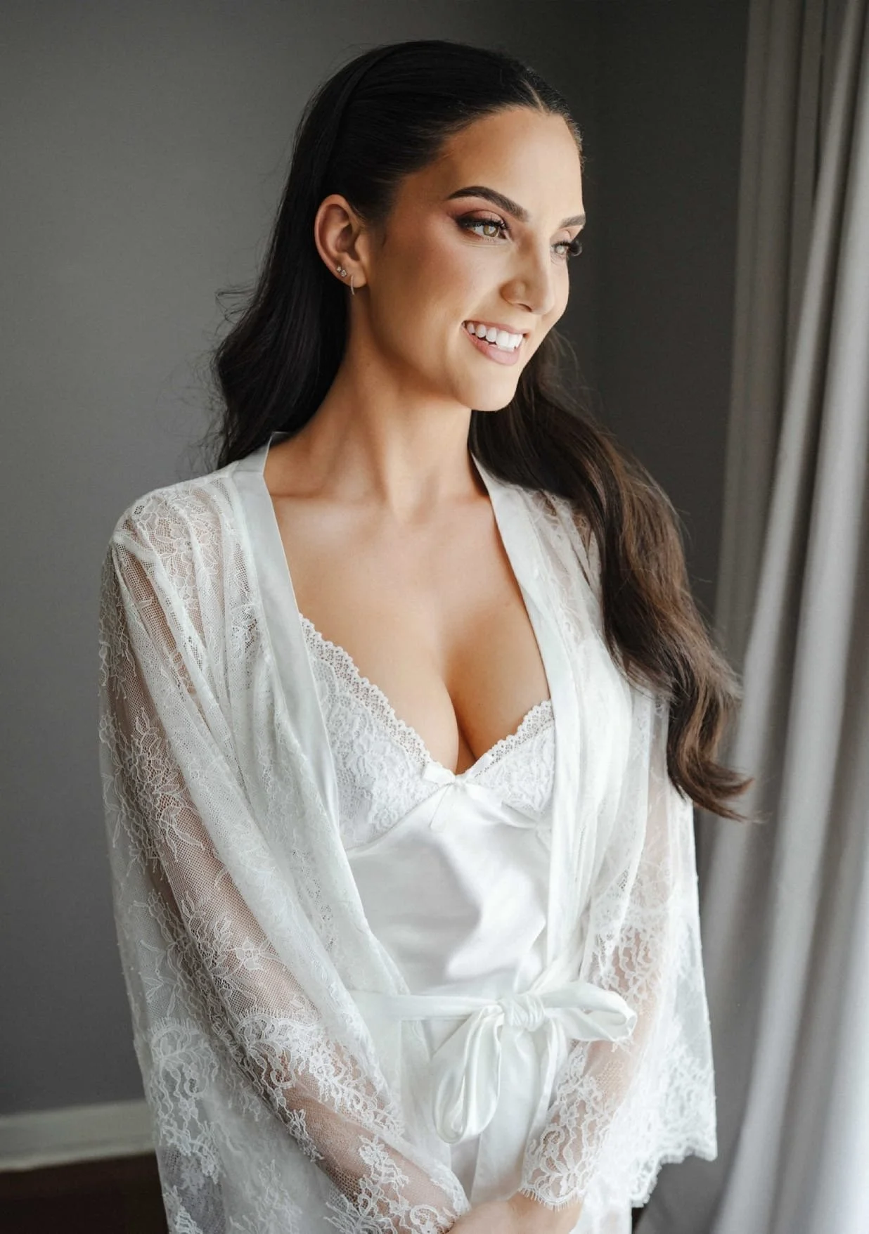 A smiling woman with long dark hair wearing white lace sleepwear, standing near a window with curtains.