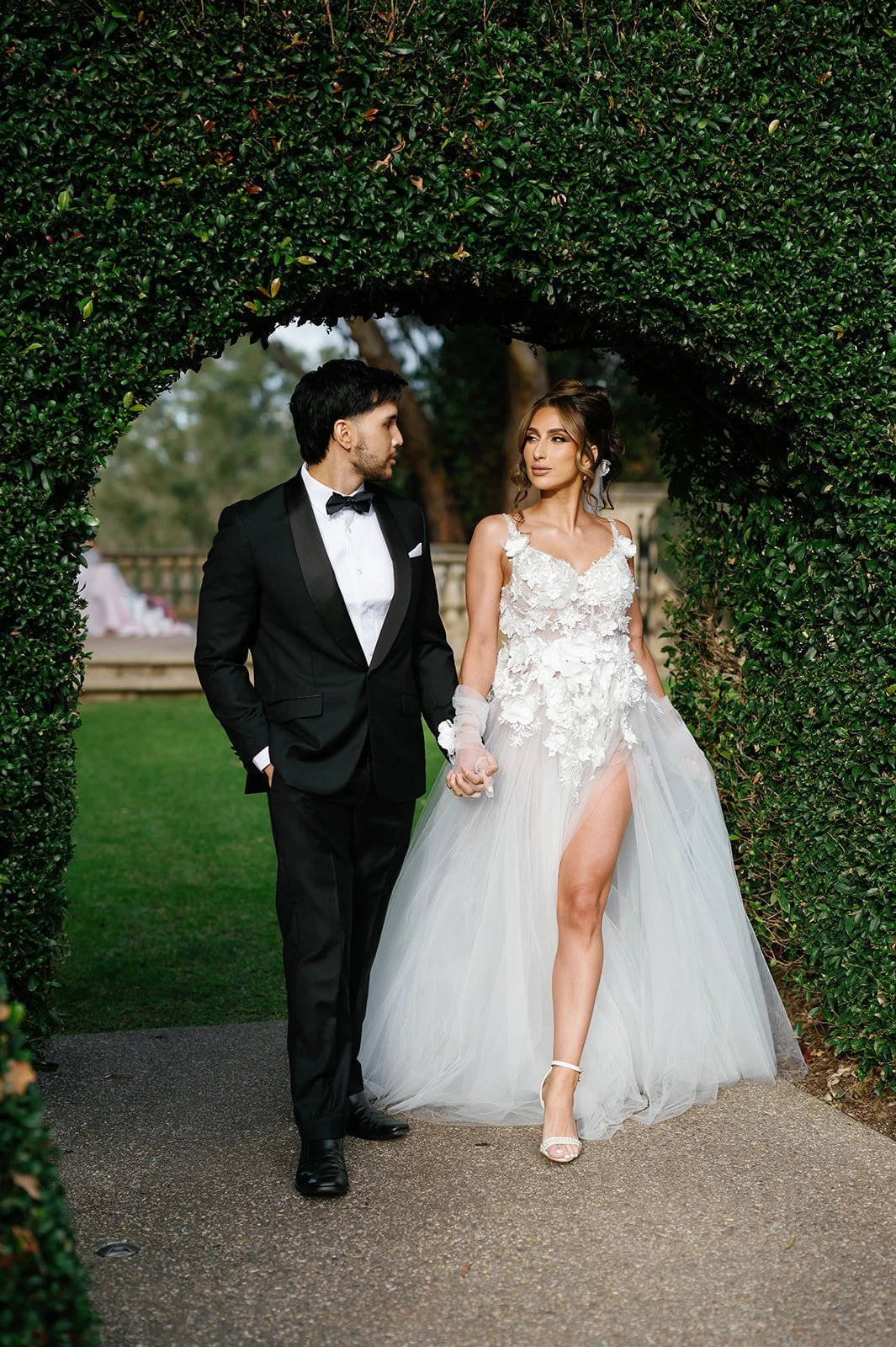 A man and woman dressed in wedding attire holding hands under a green hedge archway.