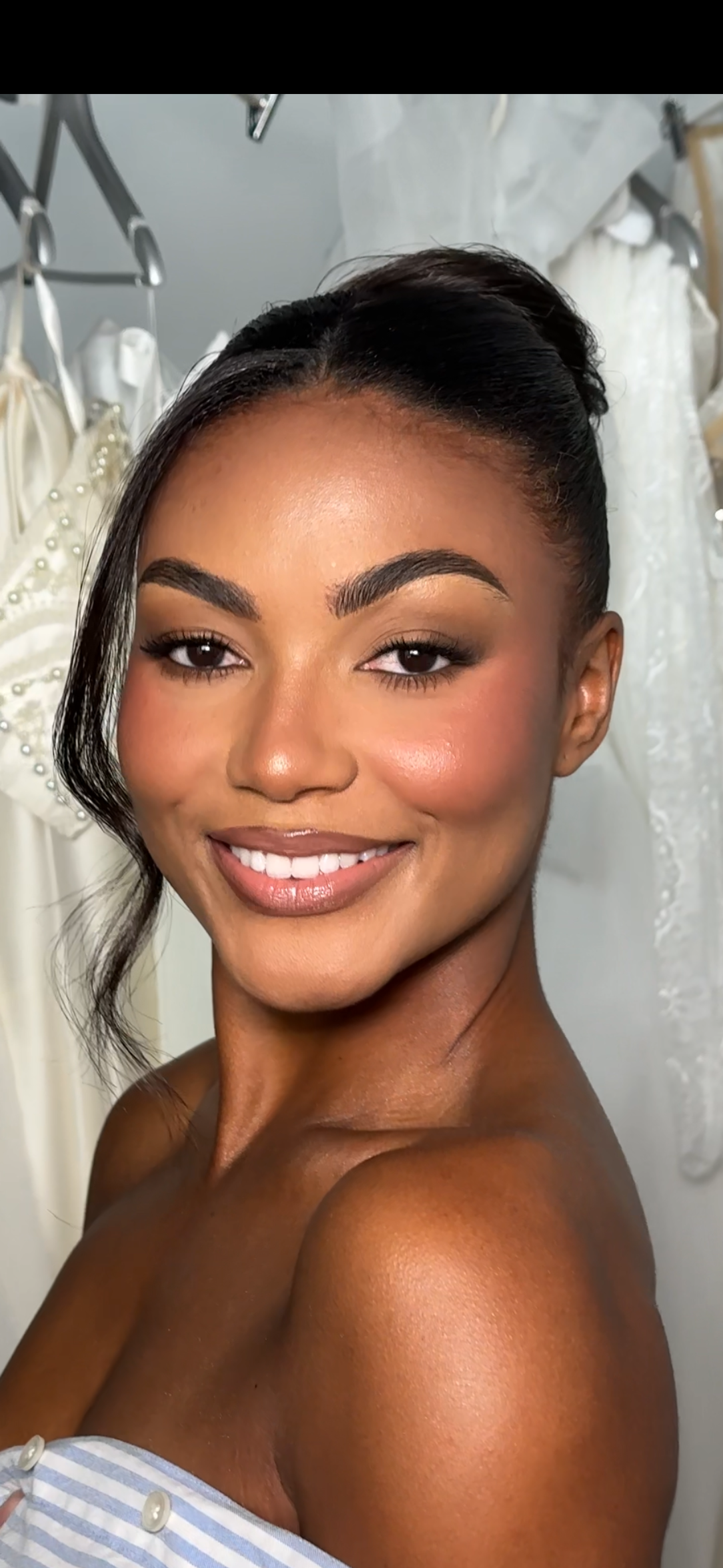 A woman with a radiant smile, makeup, and an elegant updo, inside a bridal boutique with white dresses hanging in the background.