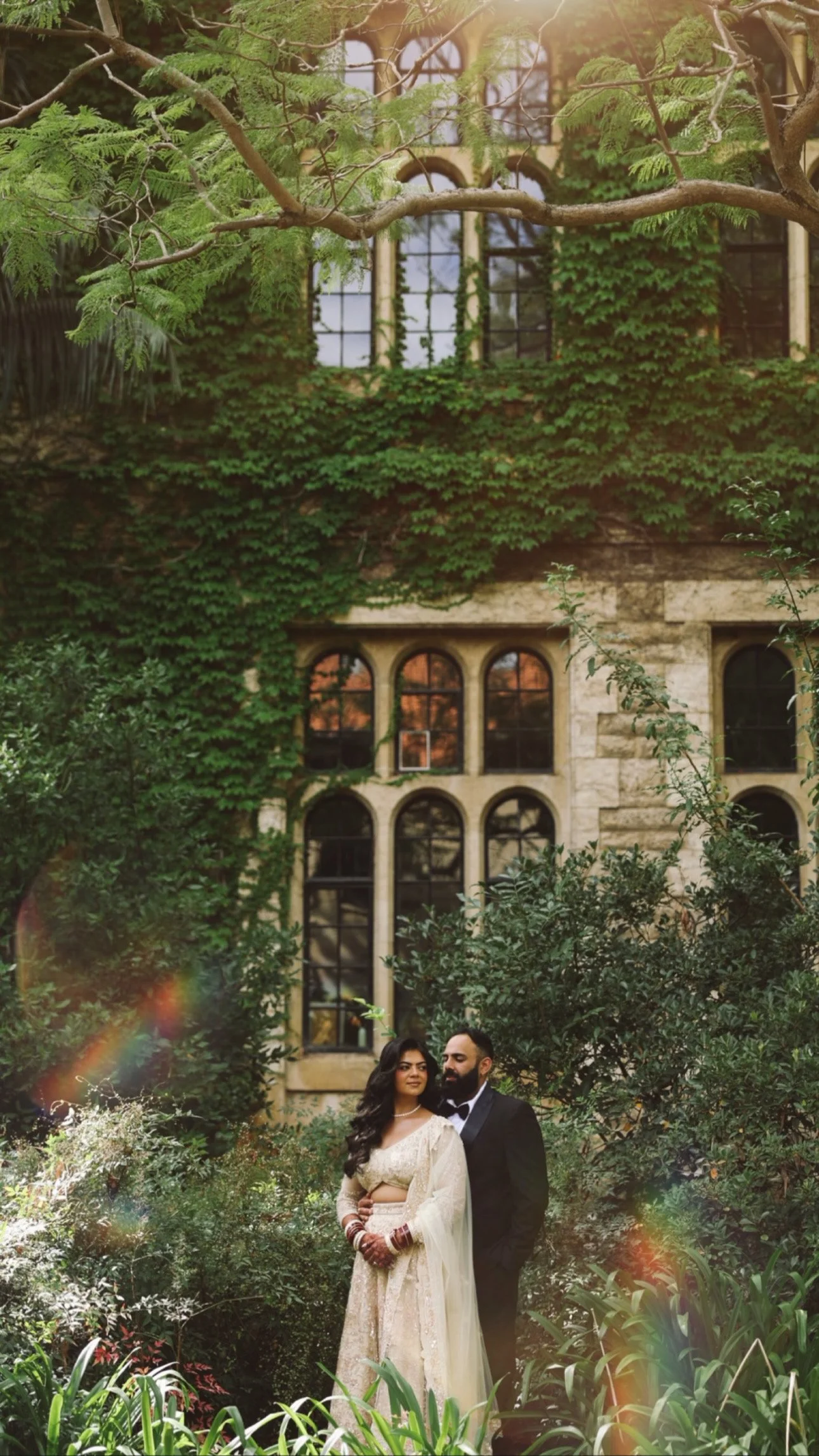 A bride and groom standing together in a lush garden, with an old stone building covered in green ivy and large arched windows in the background. The bride is wearing a traditional cream-colored dress with jewelry, and the groom is in a black suit an
