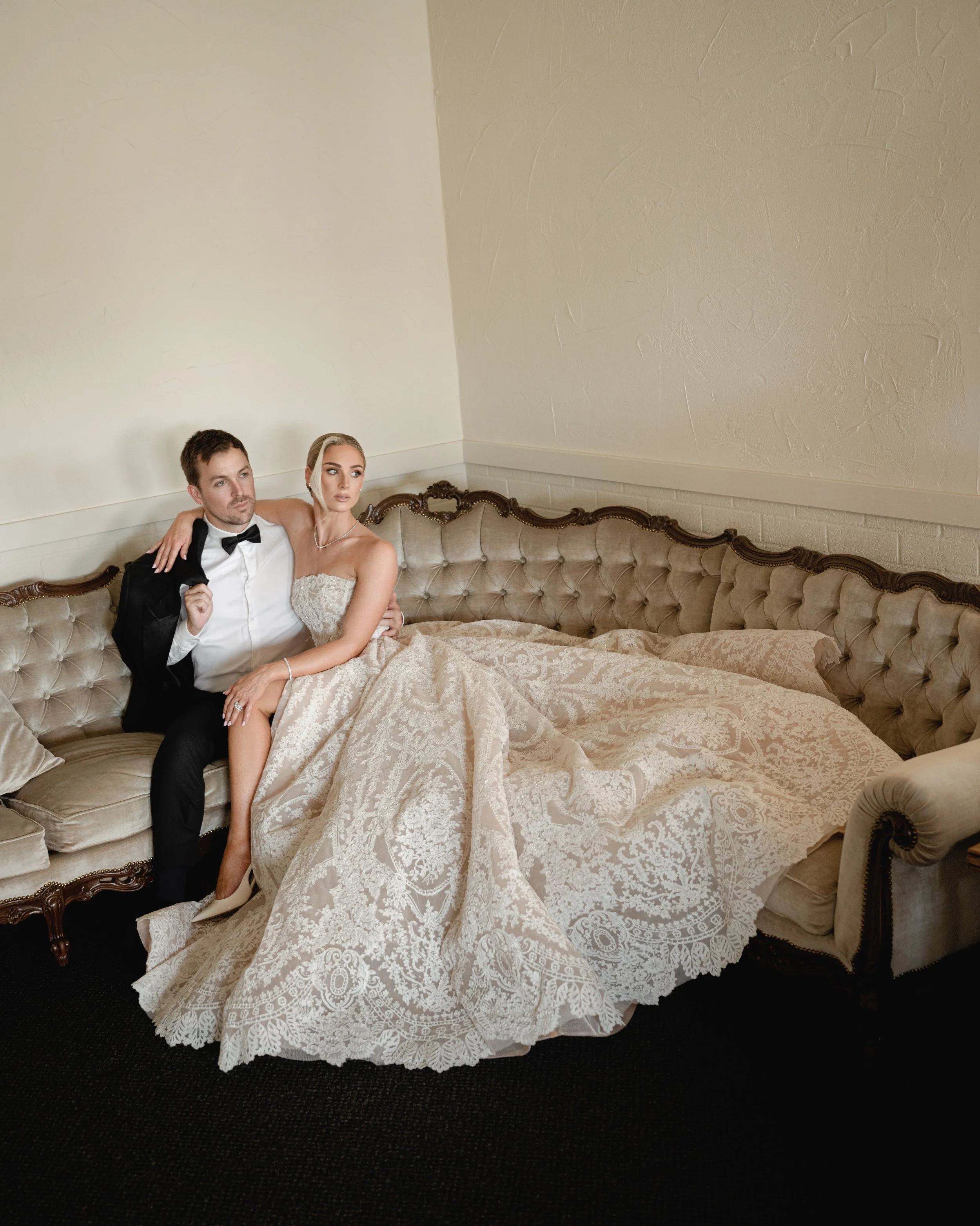 A bride and groom sitting together on a vintage beige sofa, with the bride wearing a strapless wedding gown and the groom in a tuxedo with a bow tie, in a room with cream-colored walls.