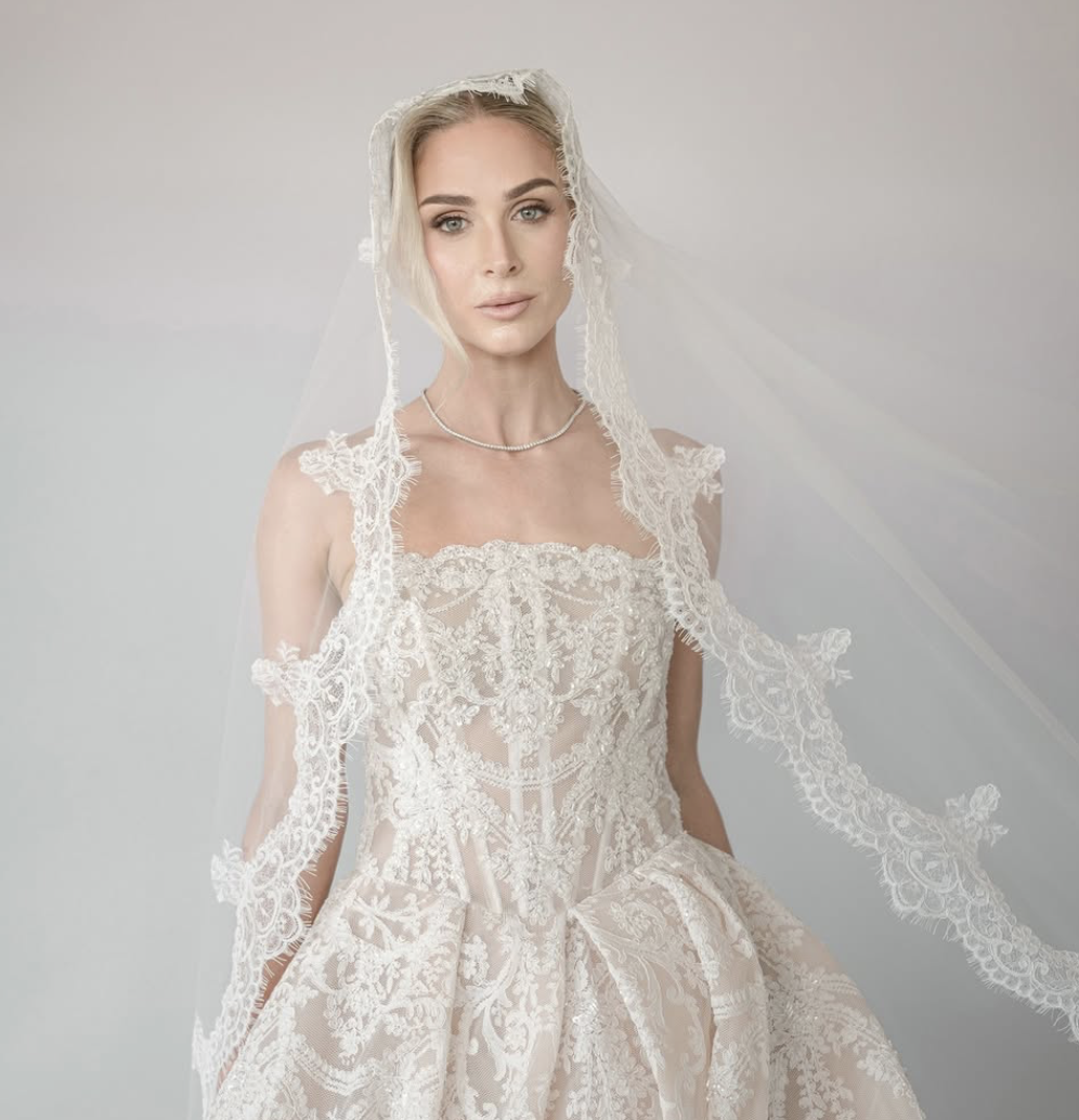 A woman wearing a wedding dress and veil, looking at the camera, against a plain background.