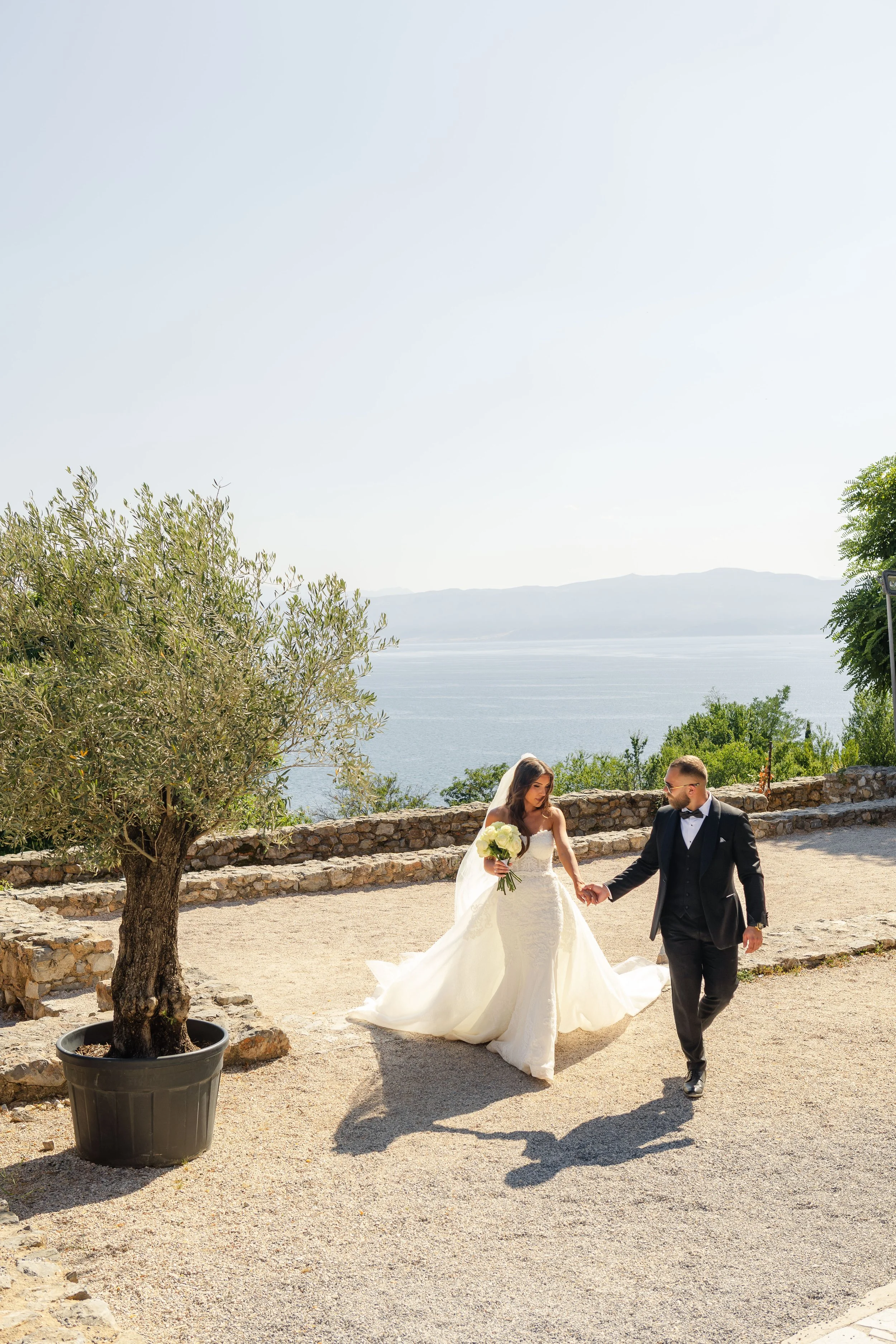 A bride and groom holding hands while walking outdoors near a body of water on a sunny day, with a tree in a pot on the left and a stone wall in the background.