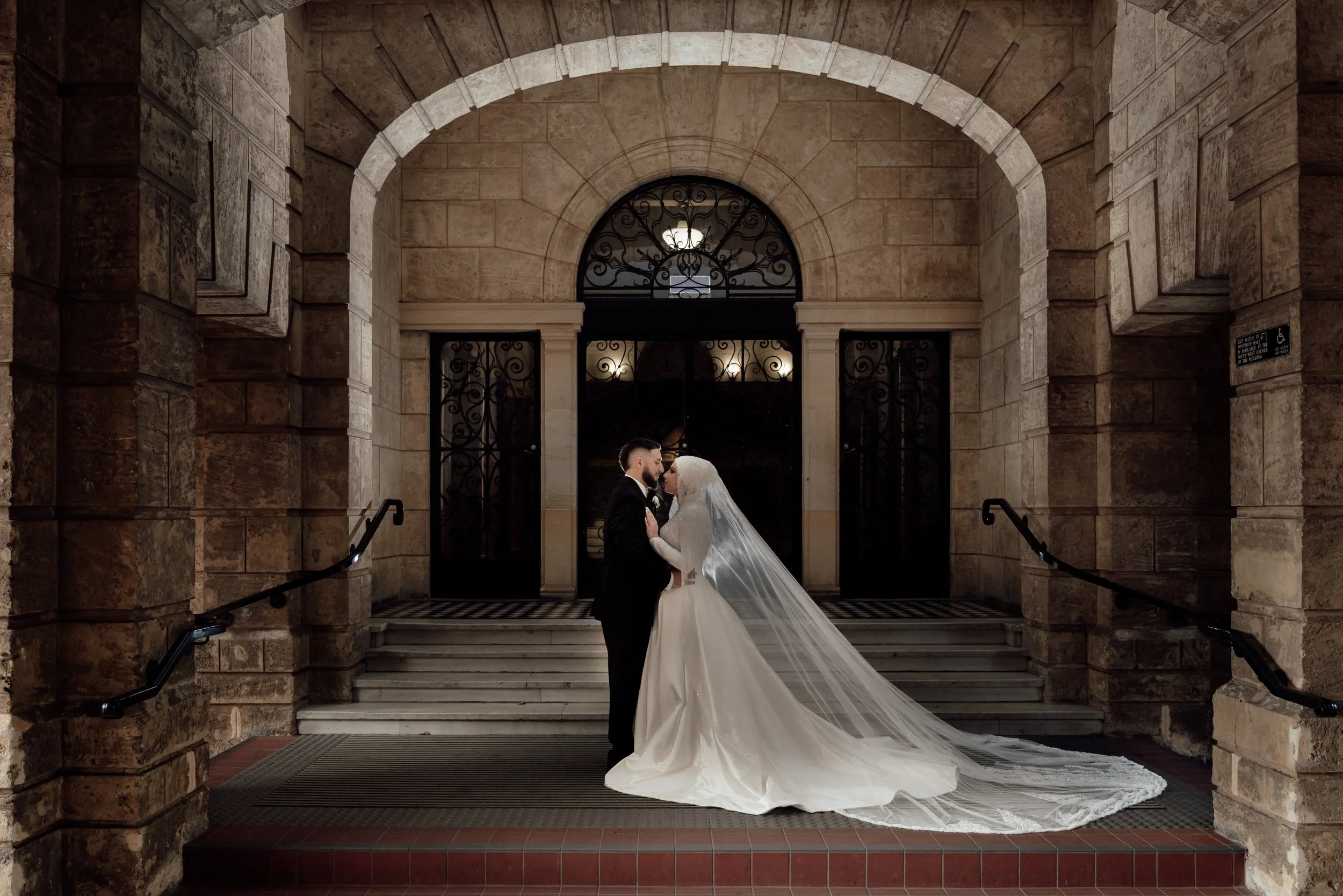 A bride and groom standing close together on the steps of a stone building, gazing into each other's eyes inside an arched doorway with ornate iron gates.