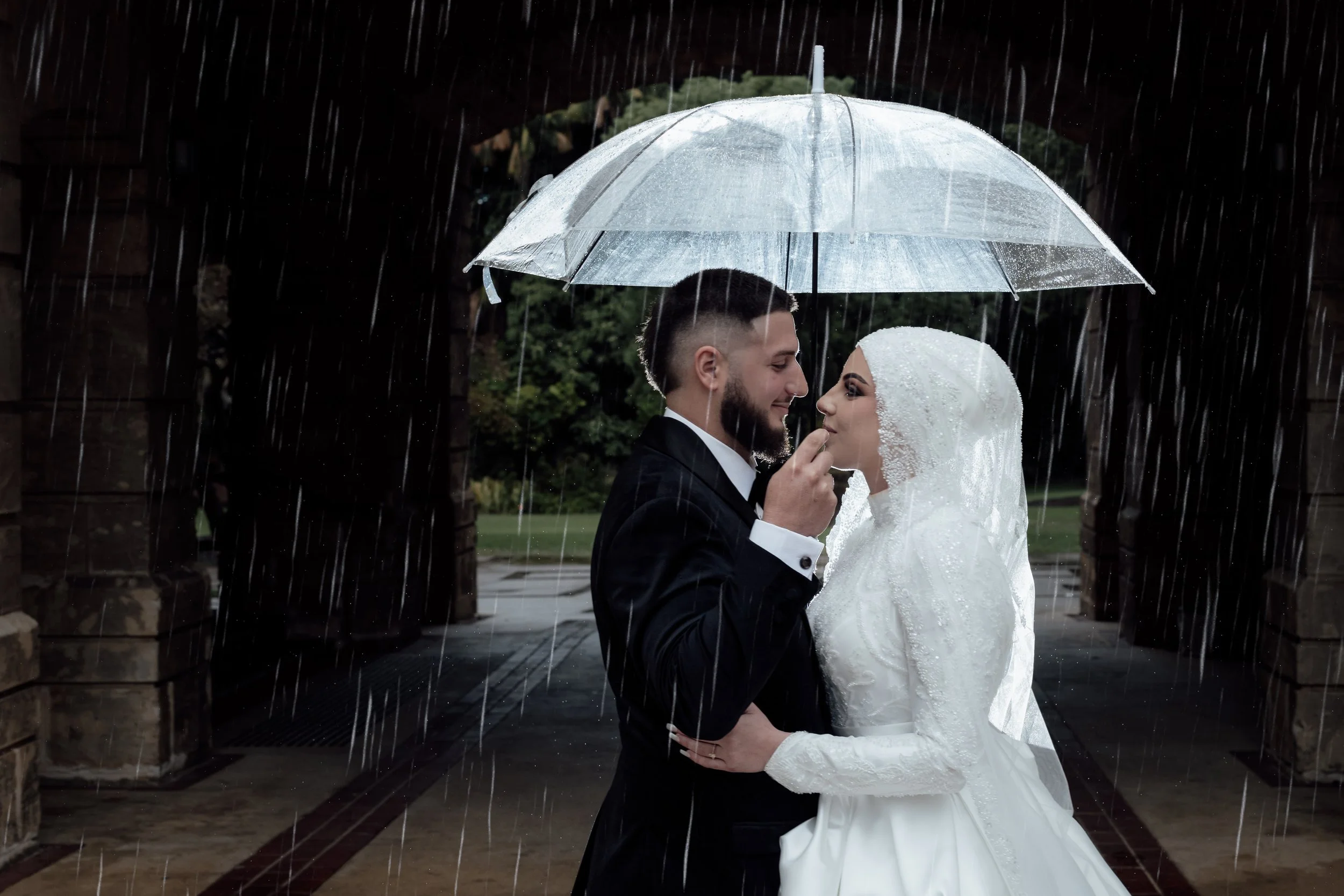 A newlywed couple stands close together under a transparent umbrella in the rain, sharing an intimate moment outdoors.