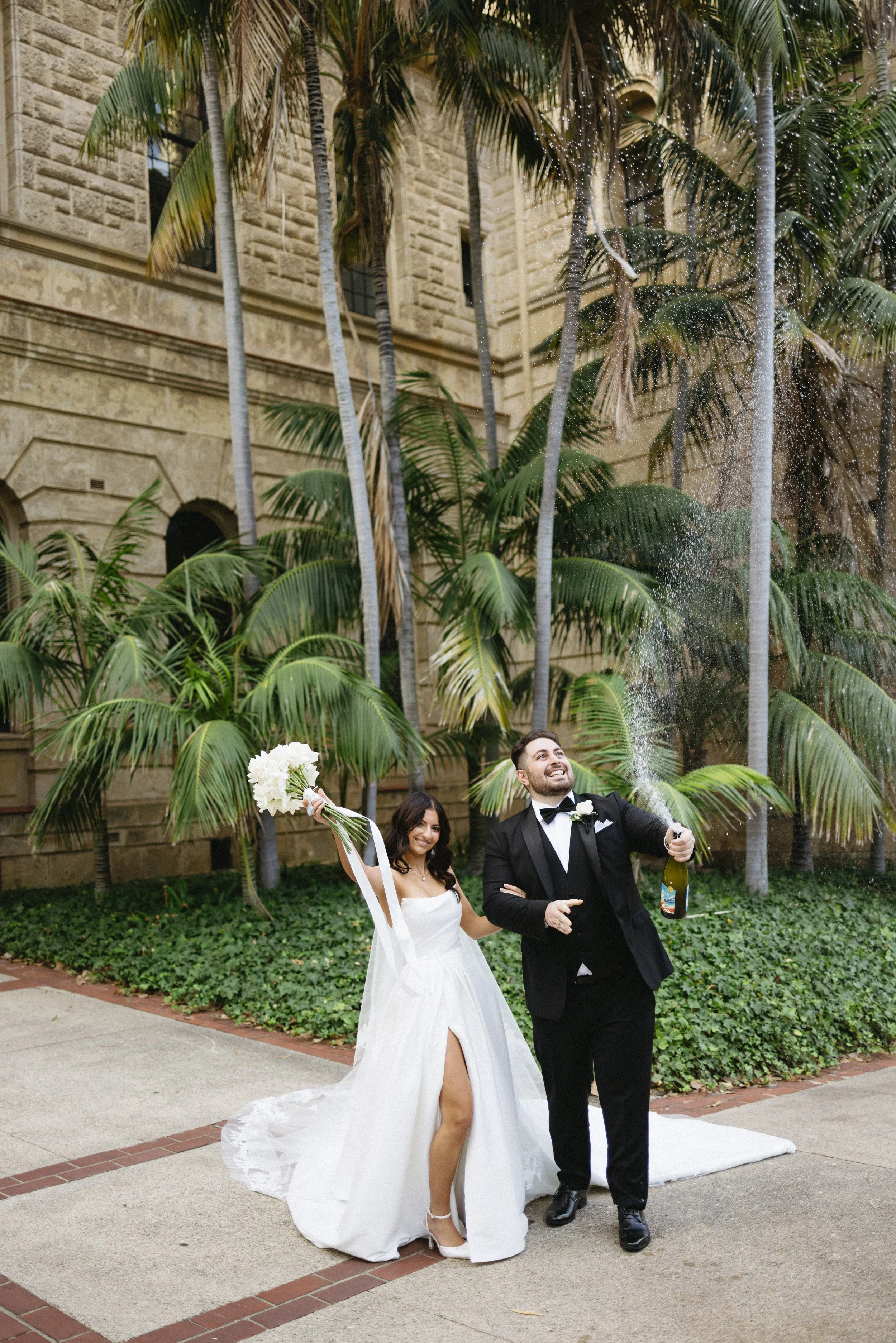 A bride and groom celebrating outdoors with champagne, palm trees, and a historic building in the background. The bride is holding a bouquet, and the groom is popping the champagne, which is spraying.