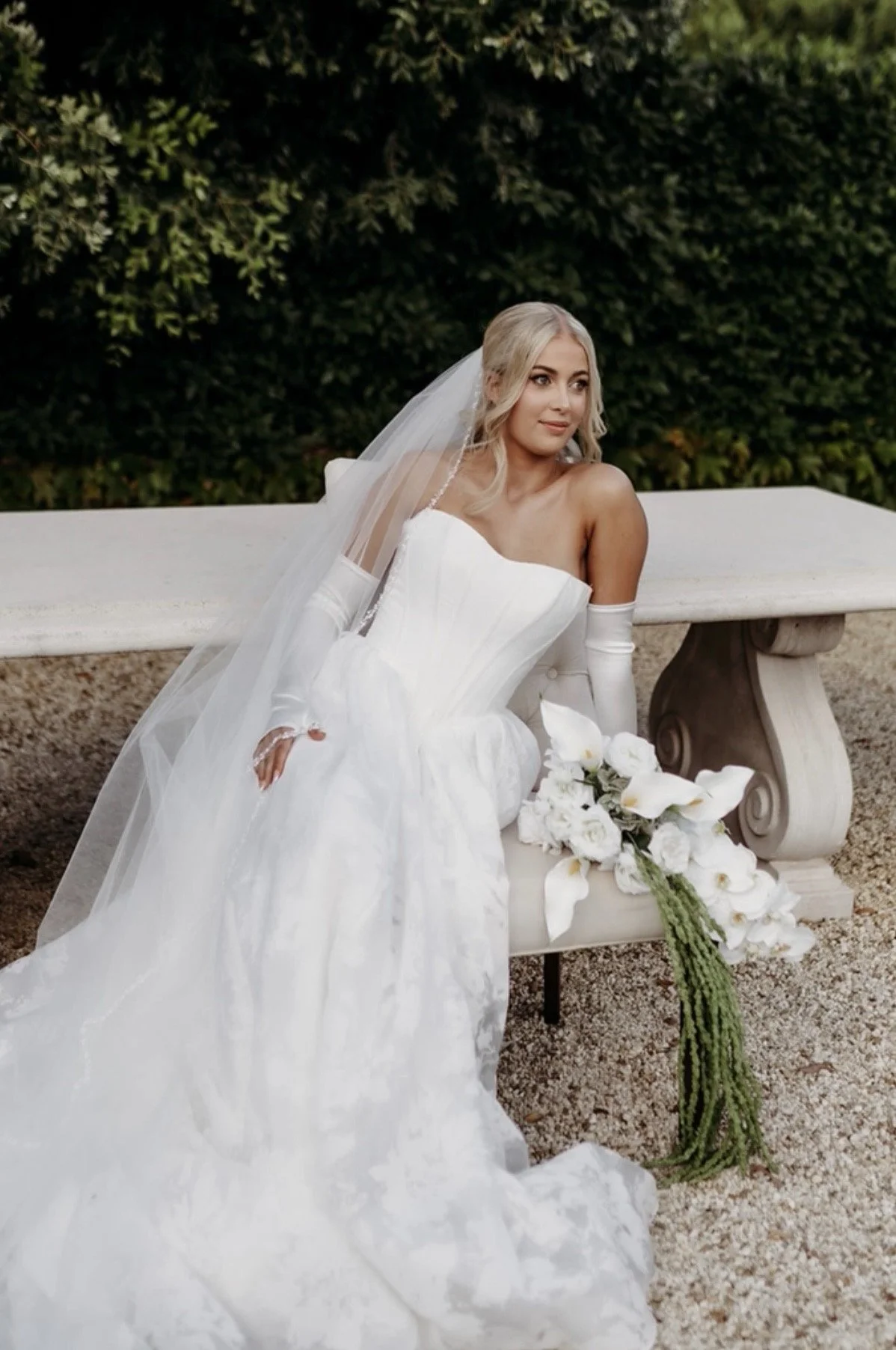 Bride in a white wedding gown and a long veil sitting on a stone bench outdoors, holding a bouquet of white calla lilies and roses with green foliage, in front of green bushes.