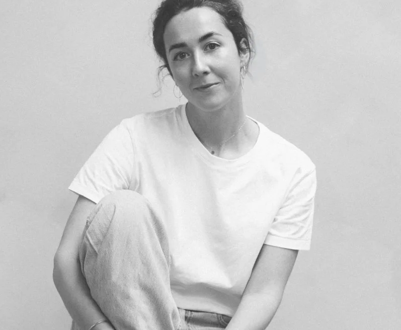 A black and white photo of a woman sitting against a plain wall, wearing a white T-shirt and light-colored pants, with her hair pulled back and a gentle smile.