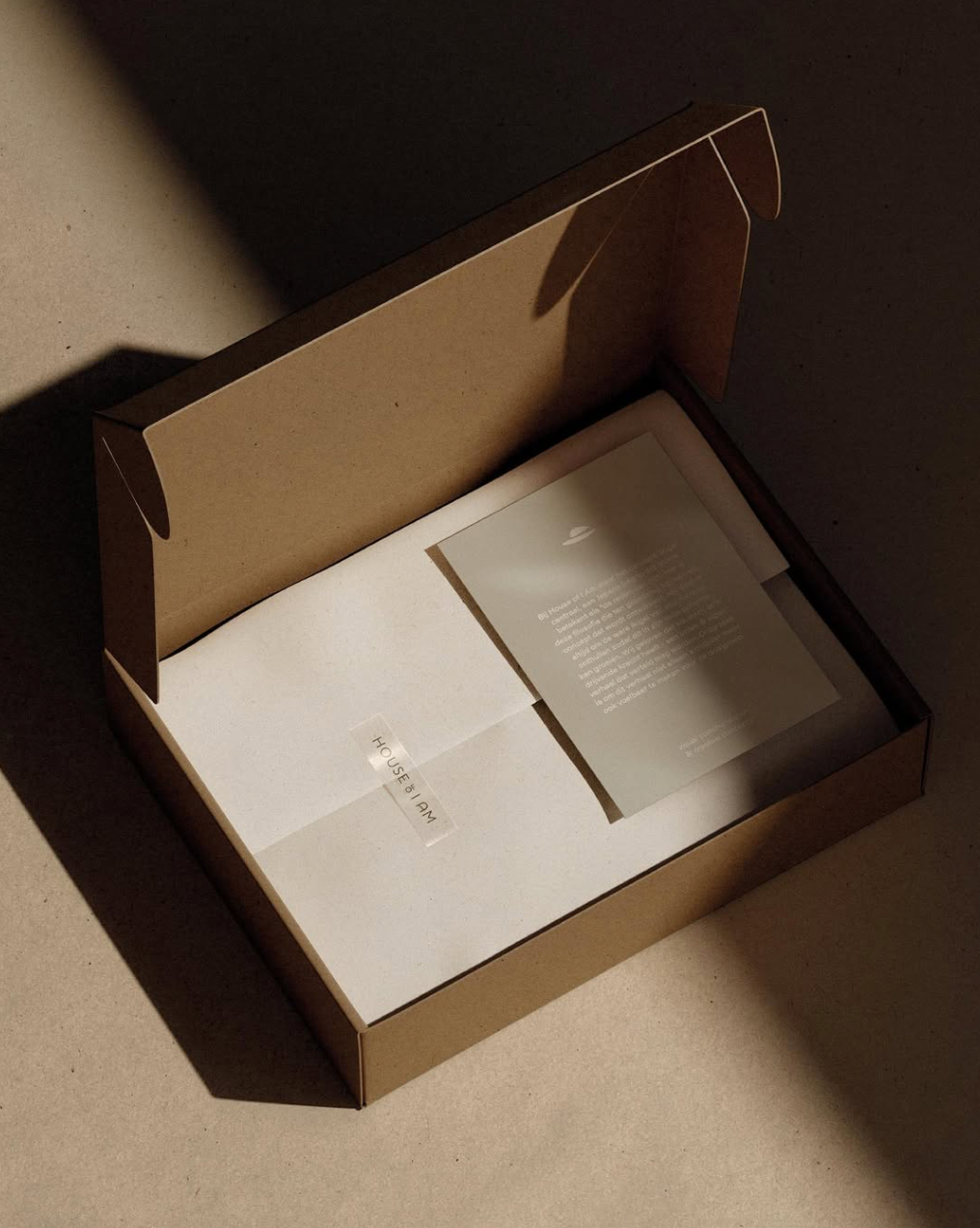 Open cardboard box with white interior containing a sheet of paper and a printed card, labeled 'HOUSE of I AM.'