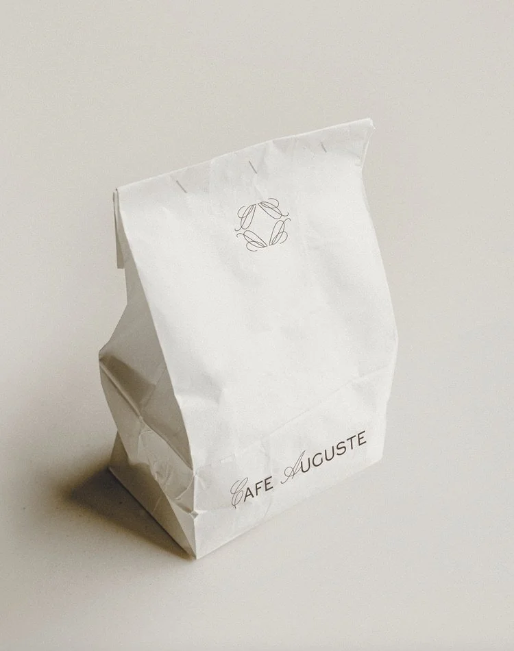 White paper bag from Cafe Auguste on a plain surface with a simple circular leaf logo and elegant lettering.