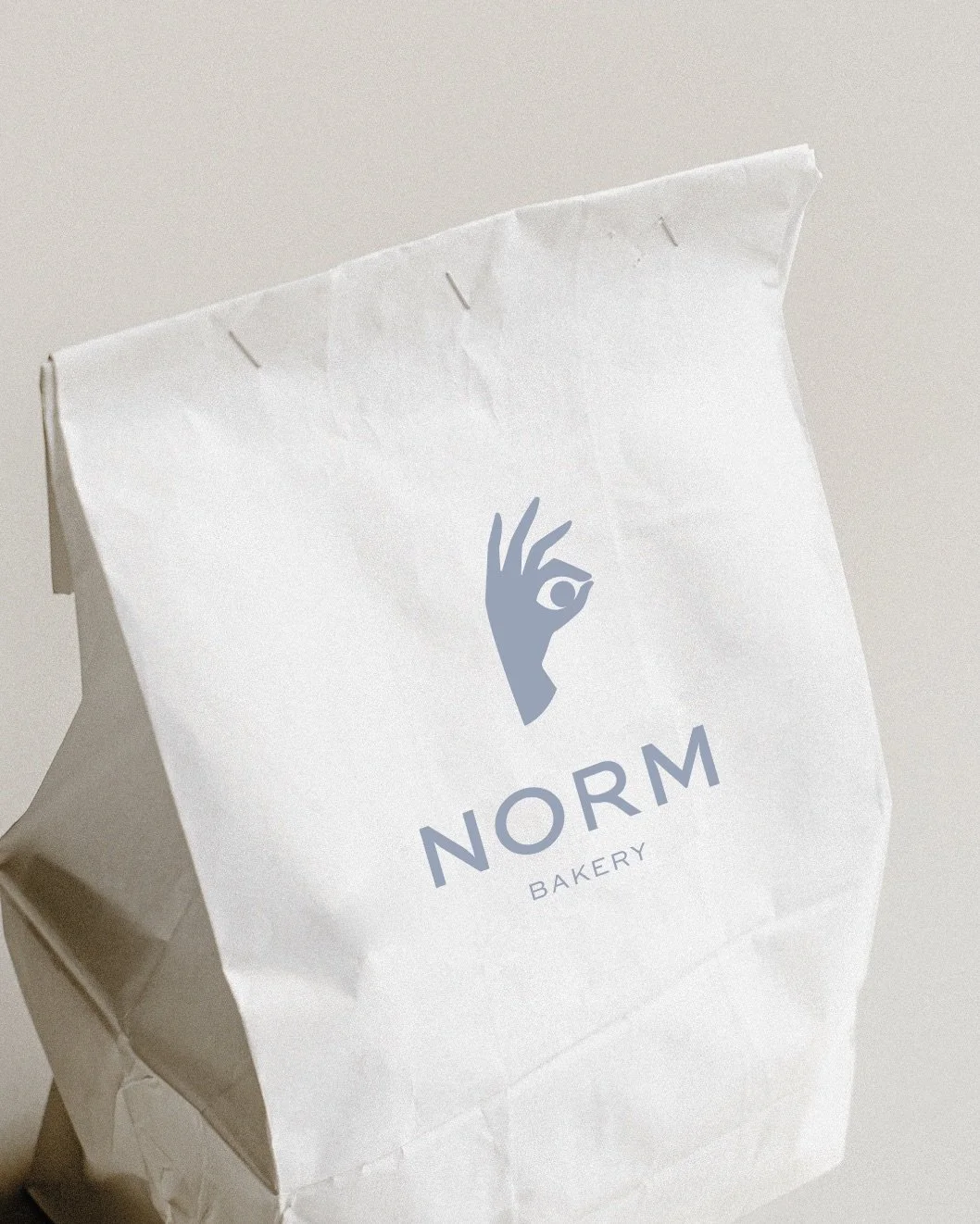 White paper bag with blue logo of a hand making an OK gesture and the words "NORM BAKERY" underneath.