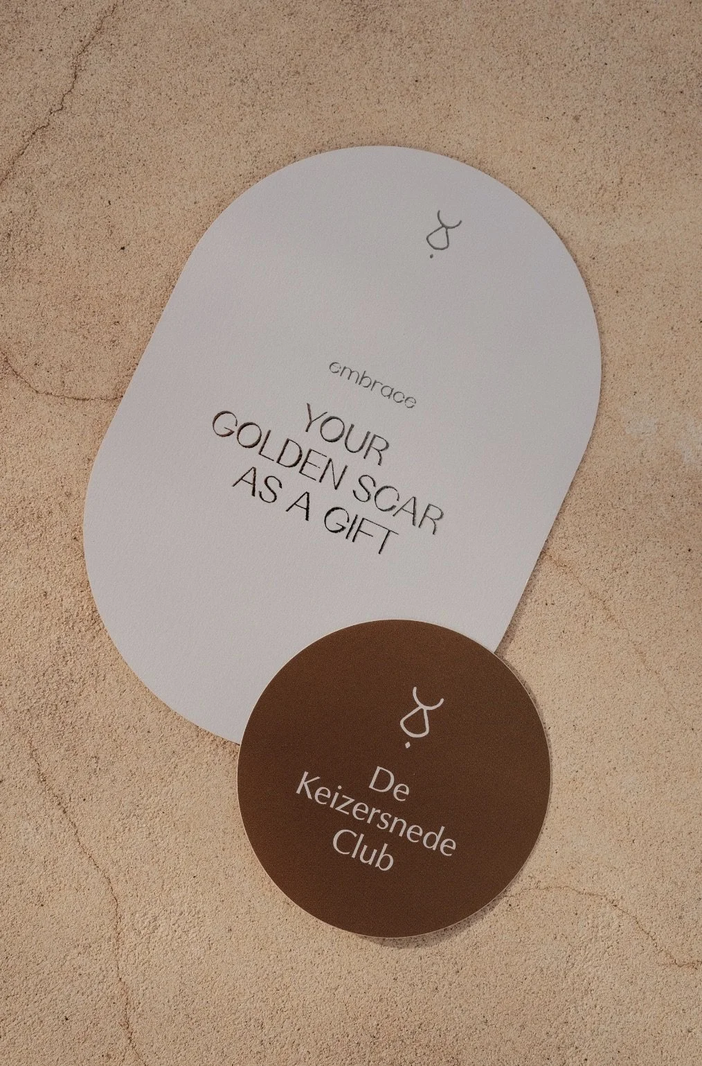 A white and a brown card on a beige textured surface. The white card reads 'embrace your golden scar as a gift,' and the brown card reads 'De Keizersnede Club' with a small stylized icon.