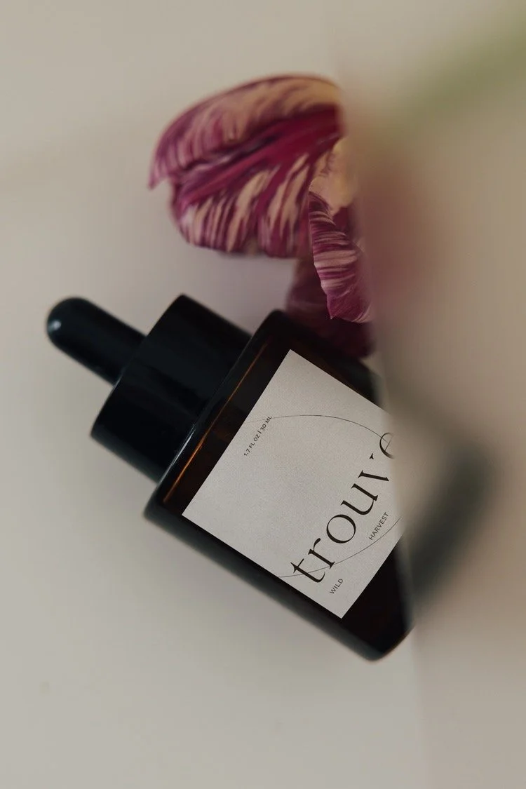 A dark amber glass bottle with a dropper cap labeled 'trouvé Wild Harvest' placed next to a pink and purple striped flower on a white surface.