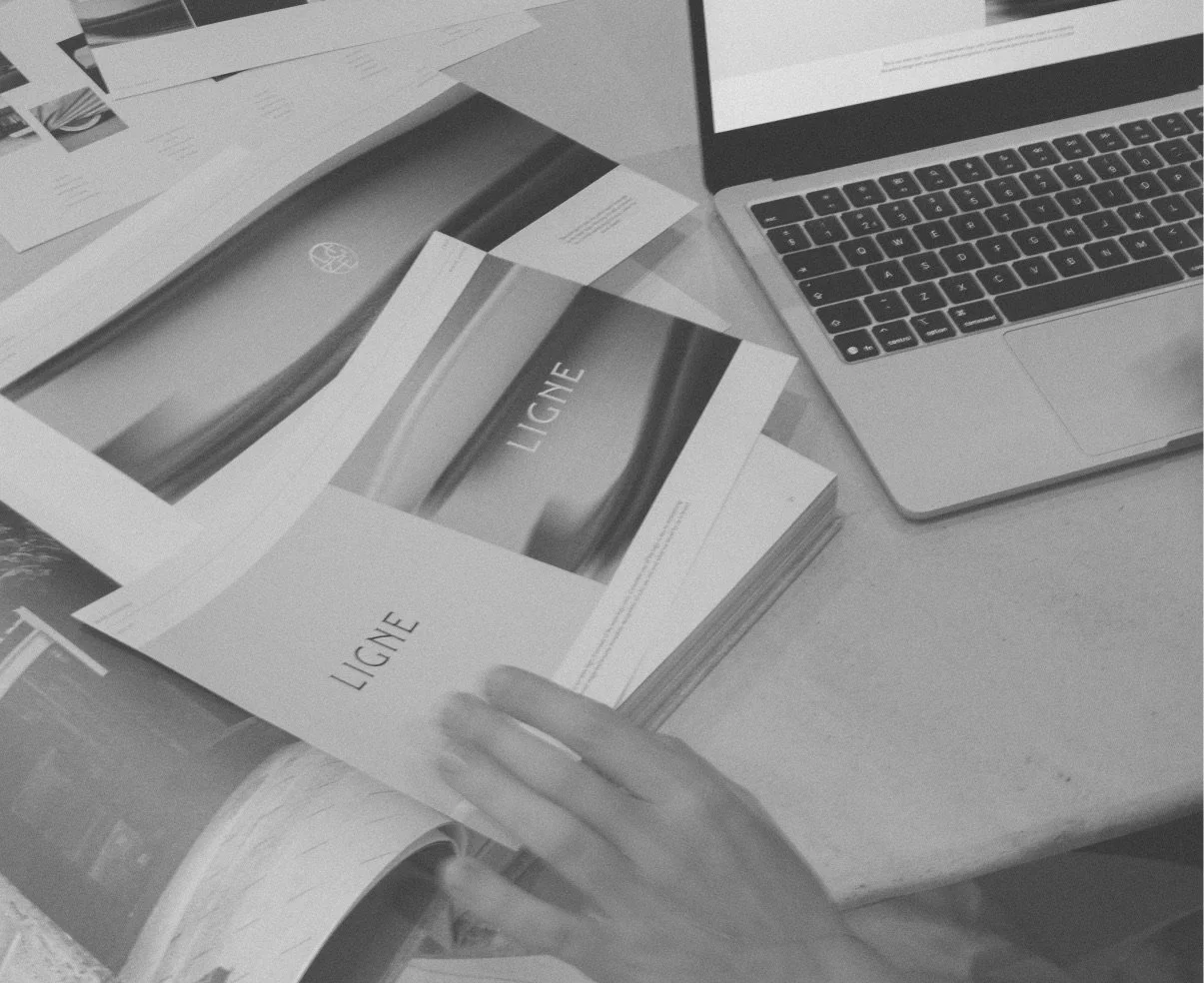 A workspace with printed brochures titled 'LIGNE', an open laptop, and scattered papers.